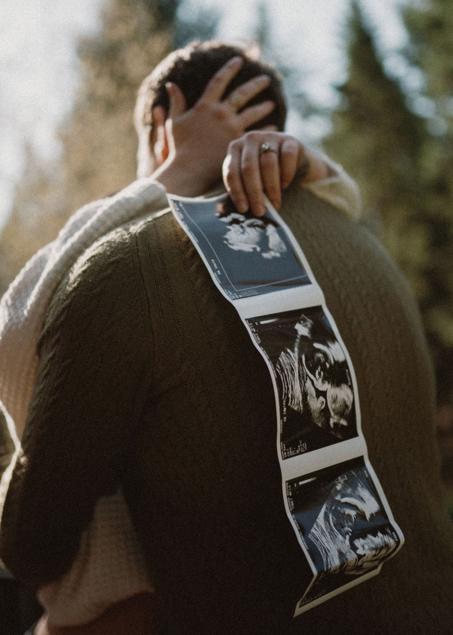 Couple embracing outdoors holding ultrasound photos, pregnancy announcement with pine trees in background.