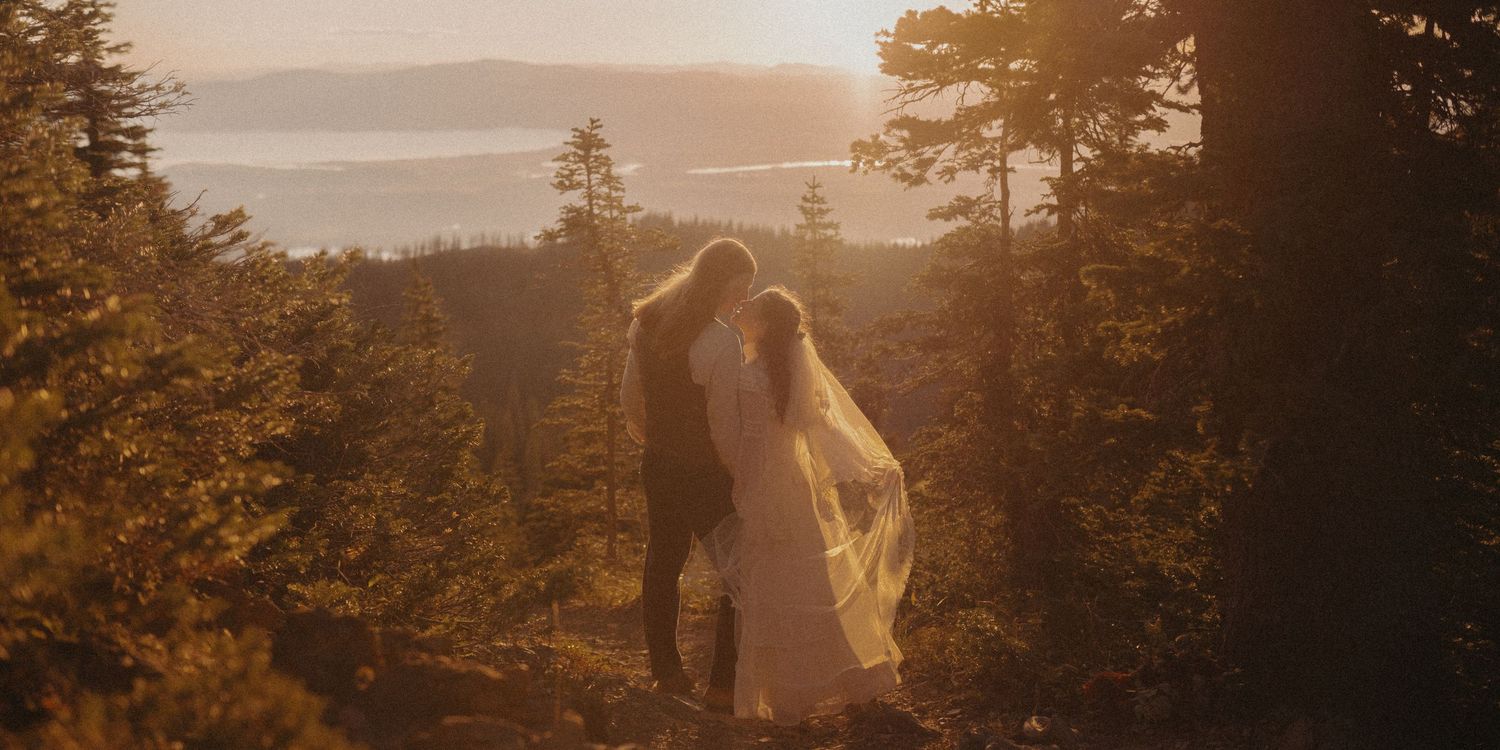 A couple embraces at sunset on a mountain overlook with golden light filtering through evergreen trees.