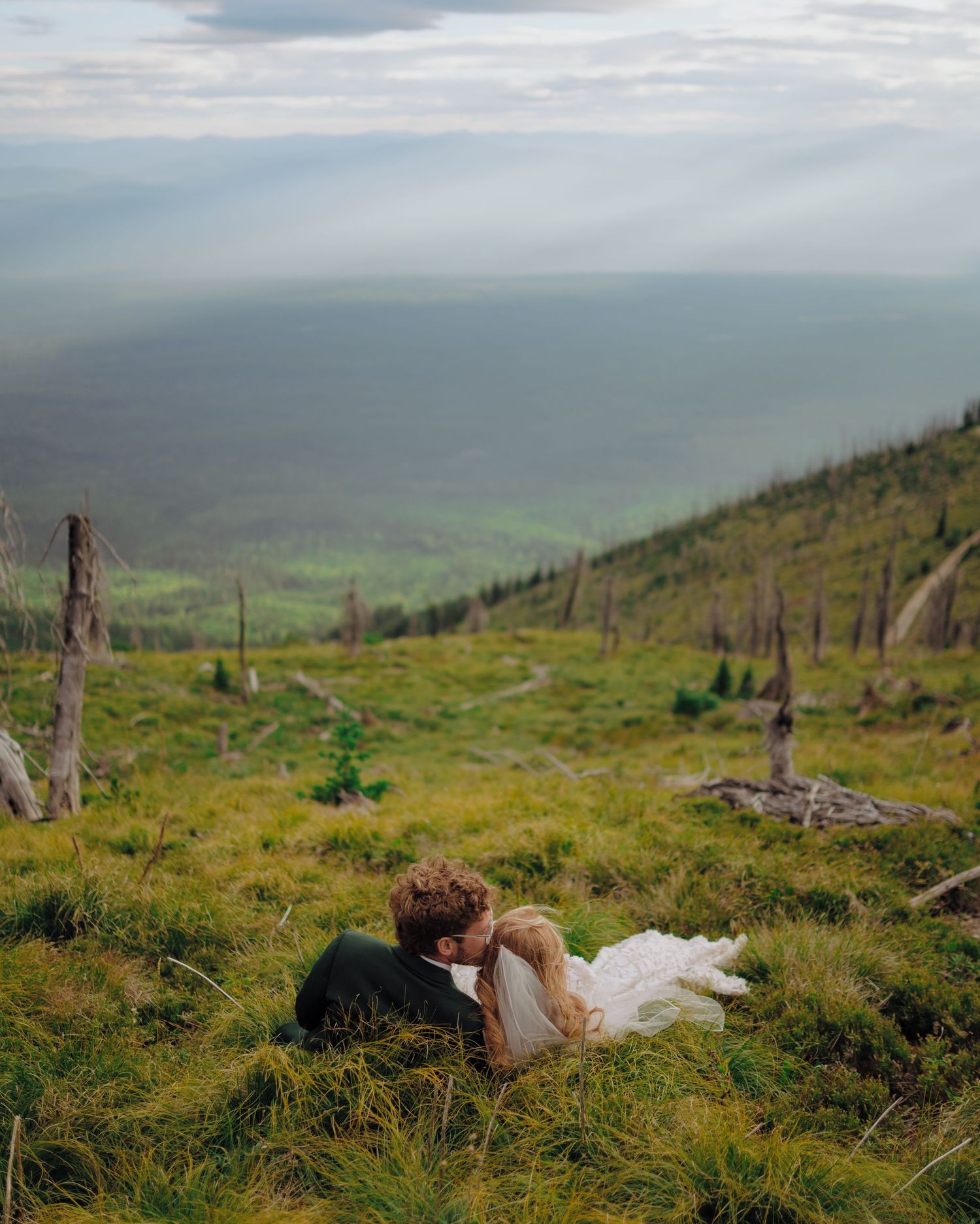 A couple embraces on a grassy hillside overlooking a misty coastal landscape under dramatic cloudy skies.