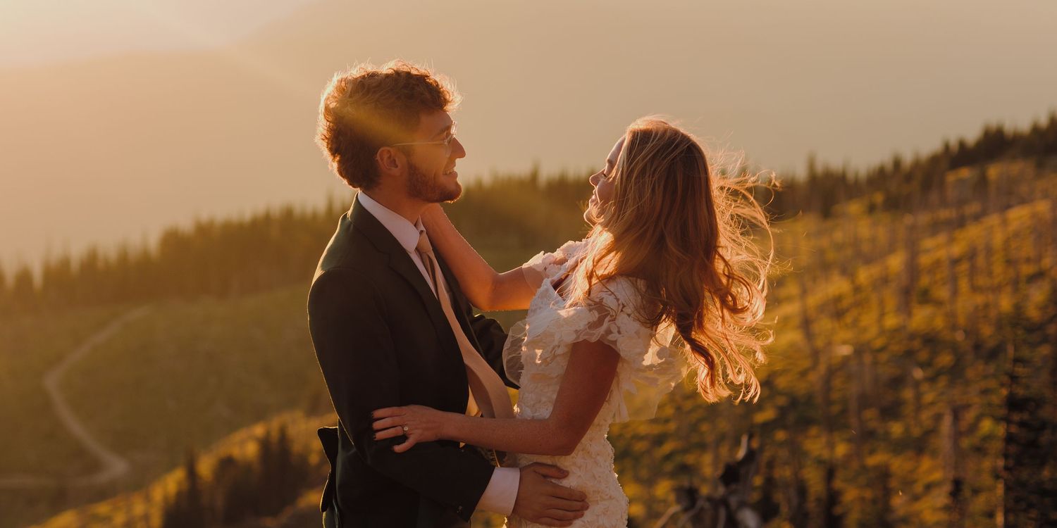 A couple embraces during golden hour on a scenic mountain overlook with sunlit evergreen trees in the background.