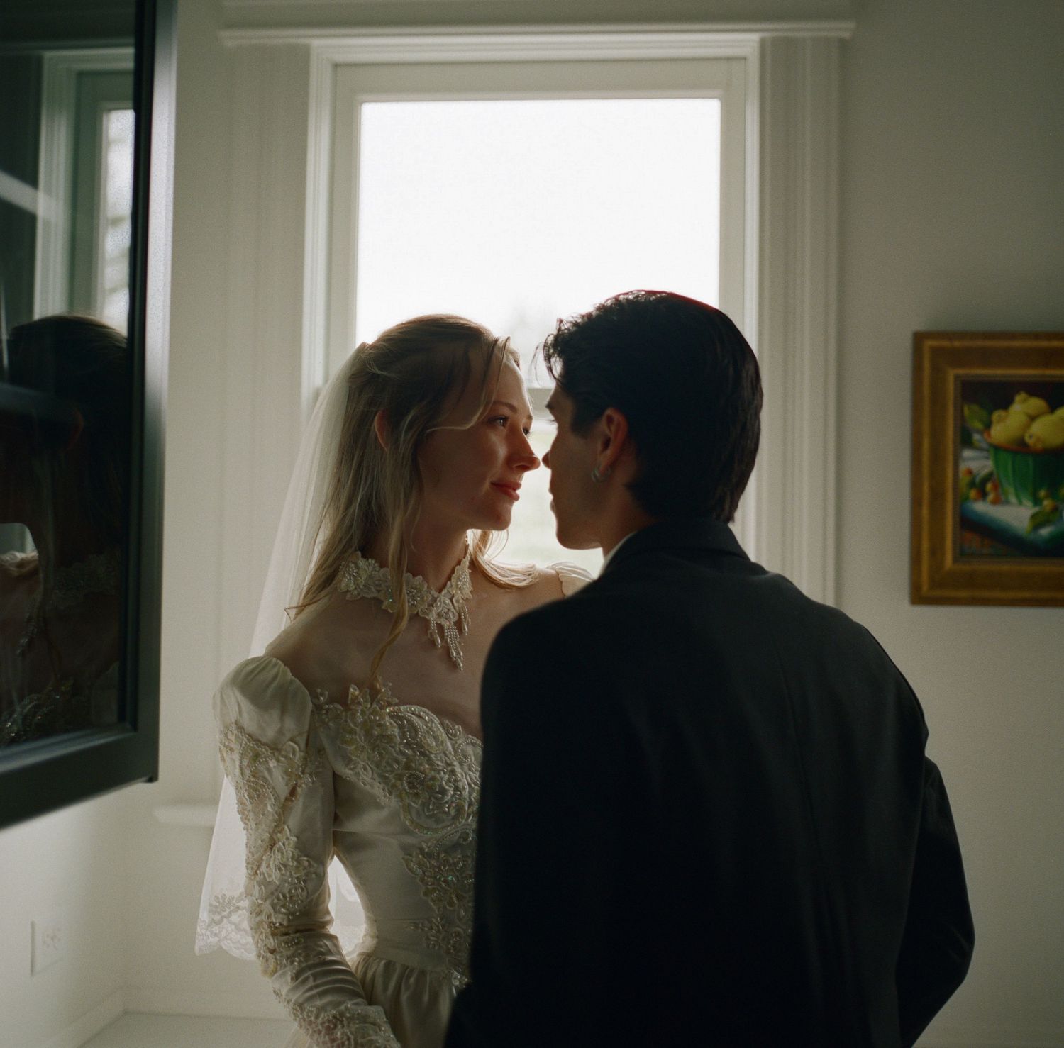 A bride in lace wedding dress and veil shares an intimate moment with groom in black suit near a sunlit window.