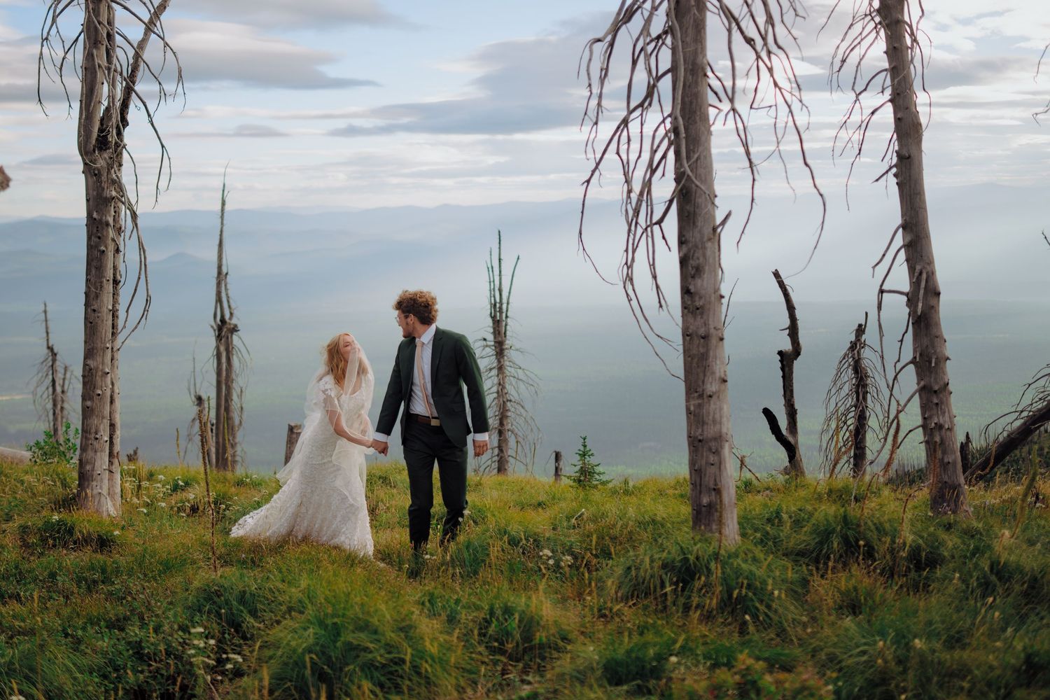 A couple walks among dead trees on a hilltop overlooking a scenic coastal view at sunset.
