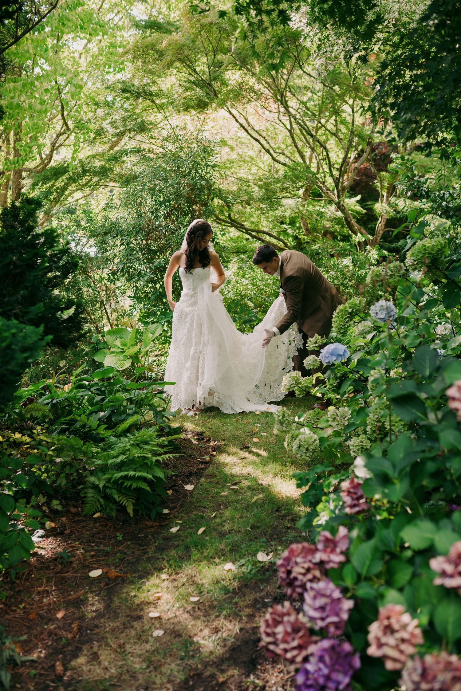 Bride and groom walk through lush garden path surrounded by ferns and hydrangeas on their wedding day.
