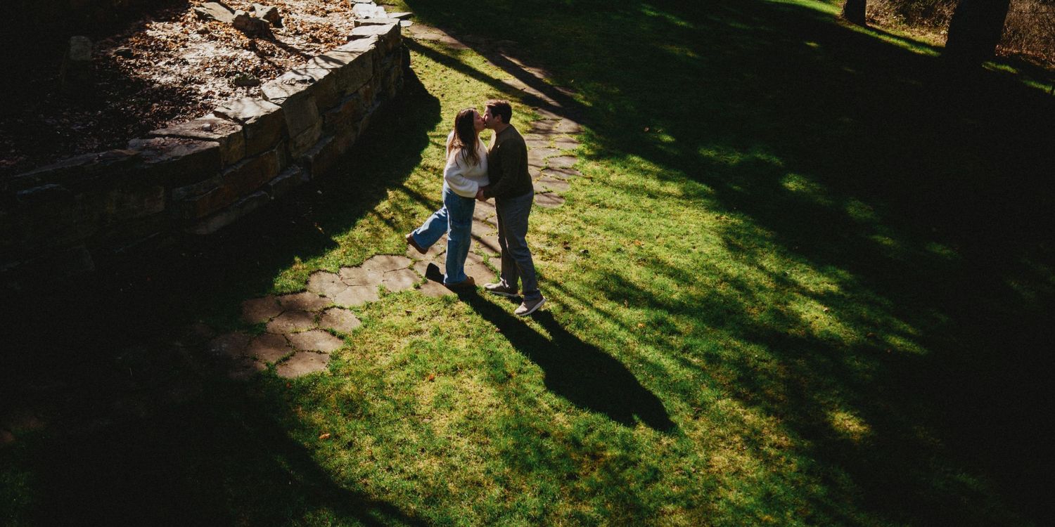 Aerial view of a romantic couple dancing on lush green grass beside a stone wall path in dappled sunlight.