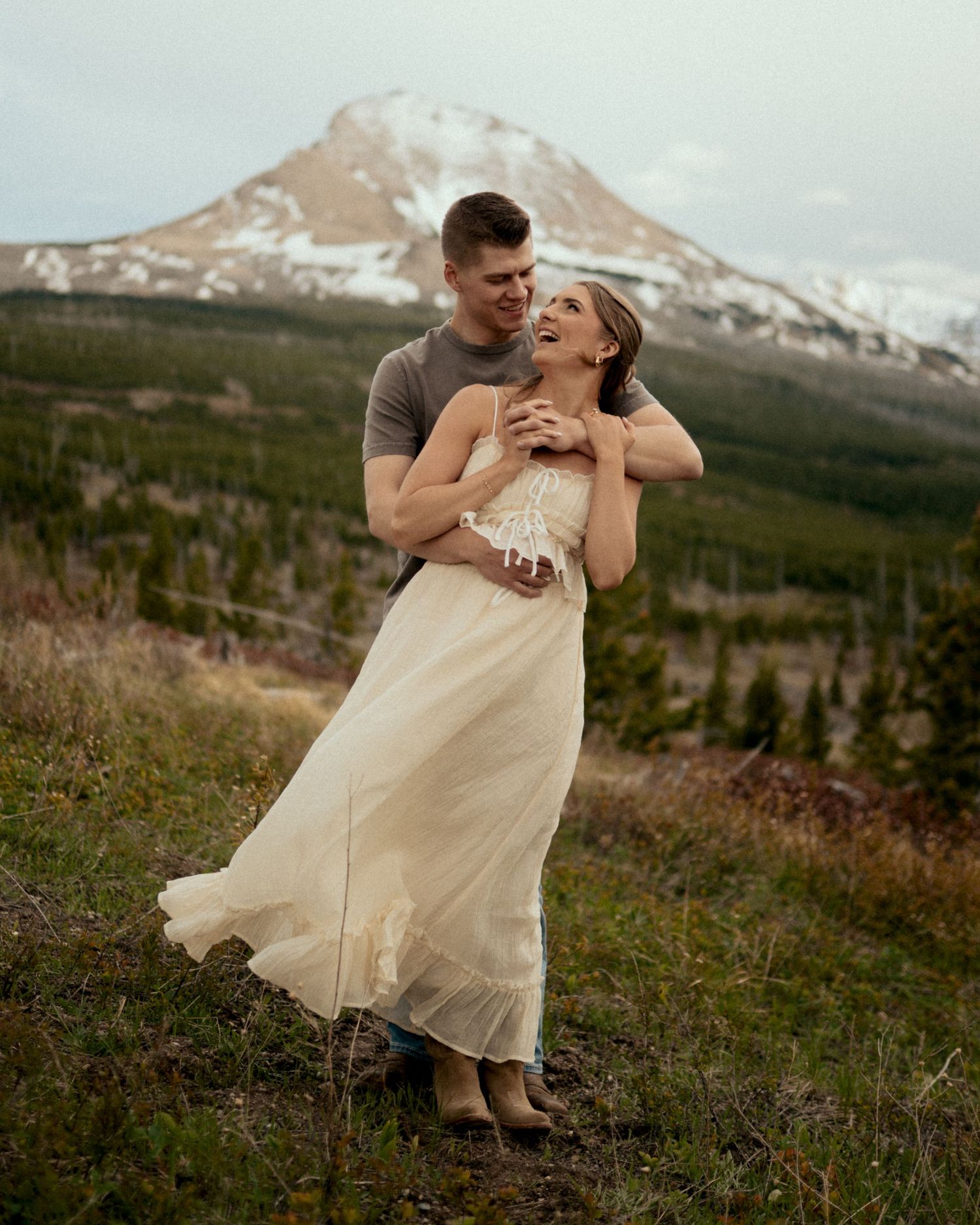 Couple embracing on a mountain meadow, woman in white dress, snow-capped peak and evergreen forest in background.