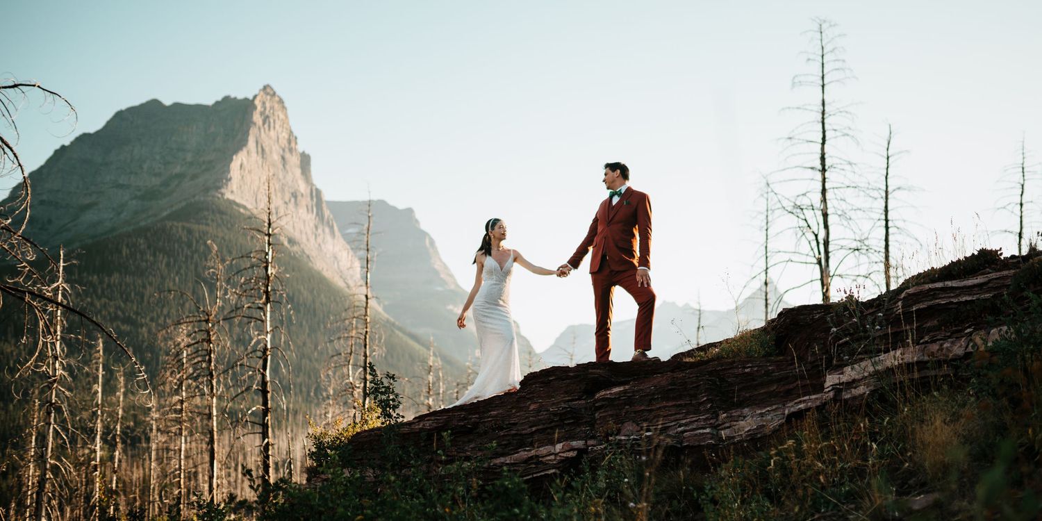 A bride and groom stand on a rocky hillside overlooking a scenic mountain landscape.