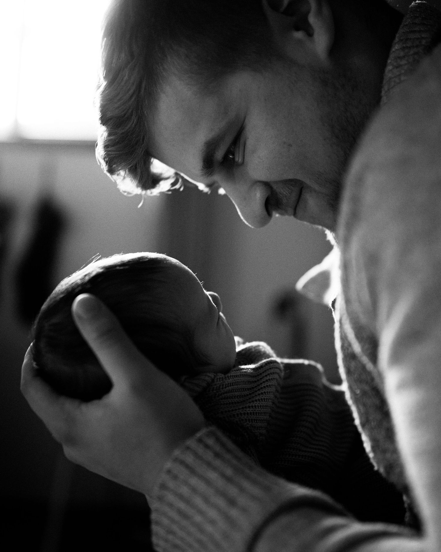 A black and white artistic photo showing a silhouette of a father smiling fondly at his newborn baby.