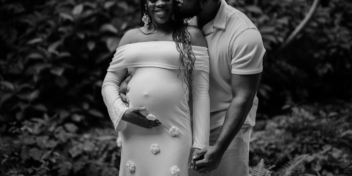 A couple shares an intimate black and white maternity photo outdoors with the pregnant woman wearing a light colored dress.