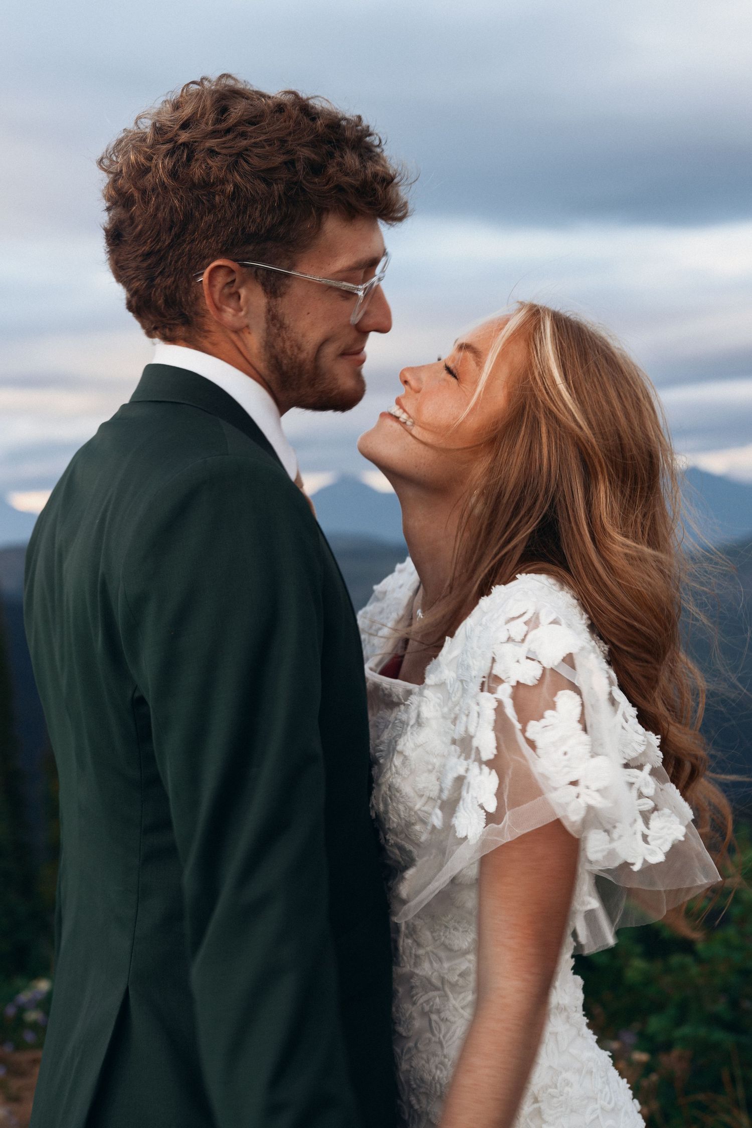 A couple in wedding attire share intimate moments against a cloudy mountain backdrop.