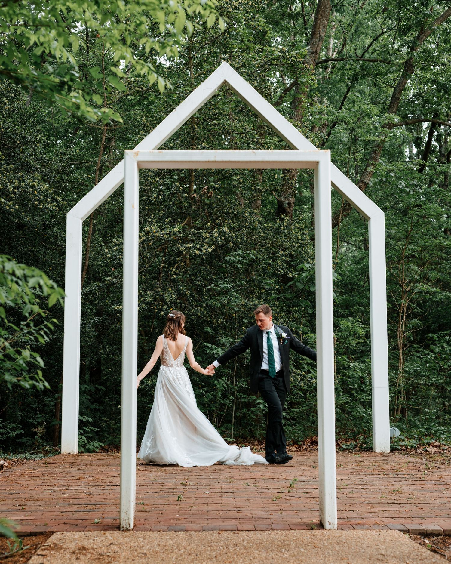 White architectural arches create wedding ceremony backdrop in wooded garden setting.