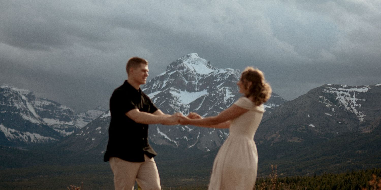 Couple holding hands dancing on a mountain overlook with dramatic stormy skies and snow-capped peaks behind them.