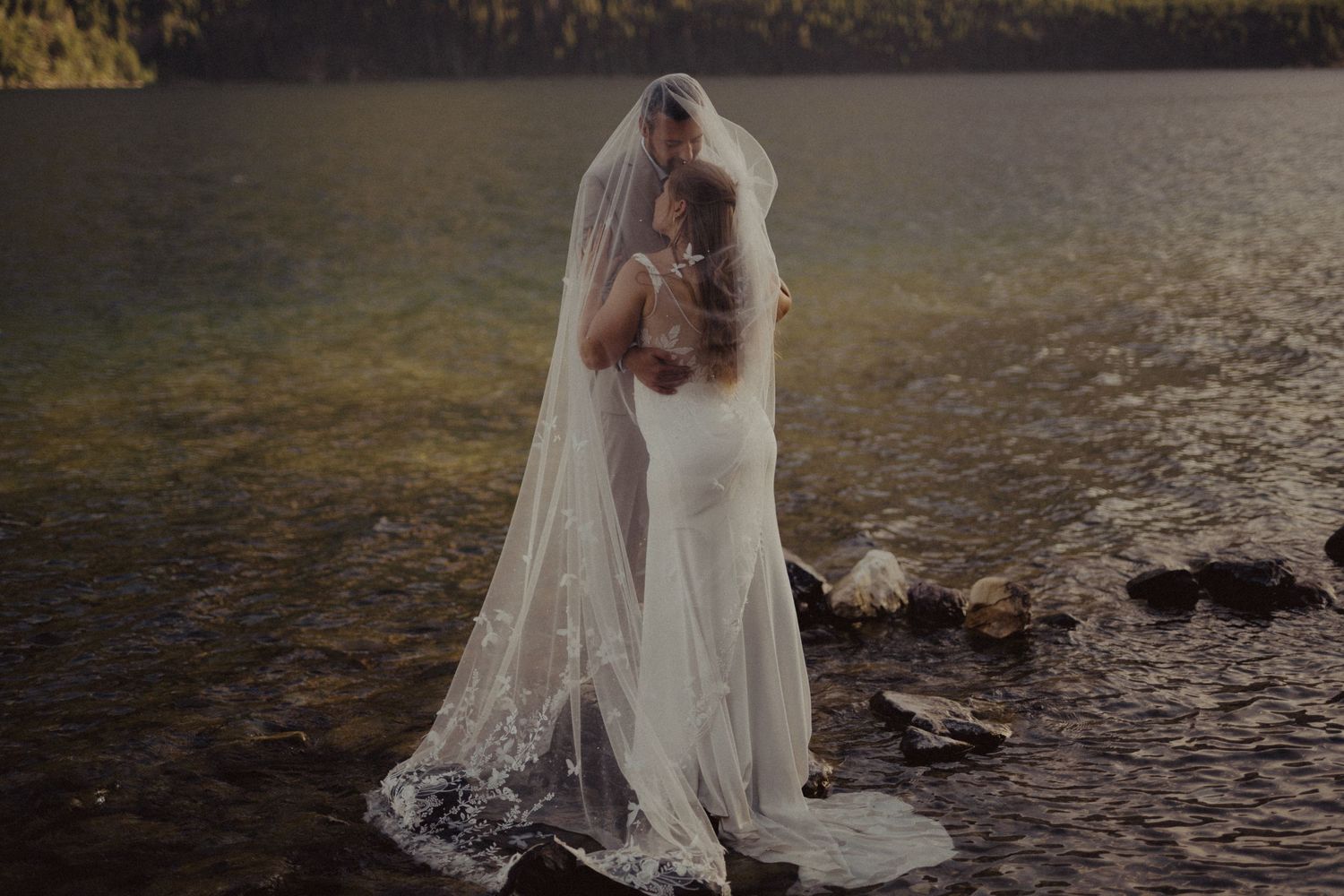 A bride in a flowing white dress walks along a scenic riverbank during a romantic outdoor photoshoot.