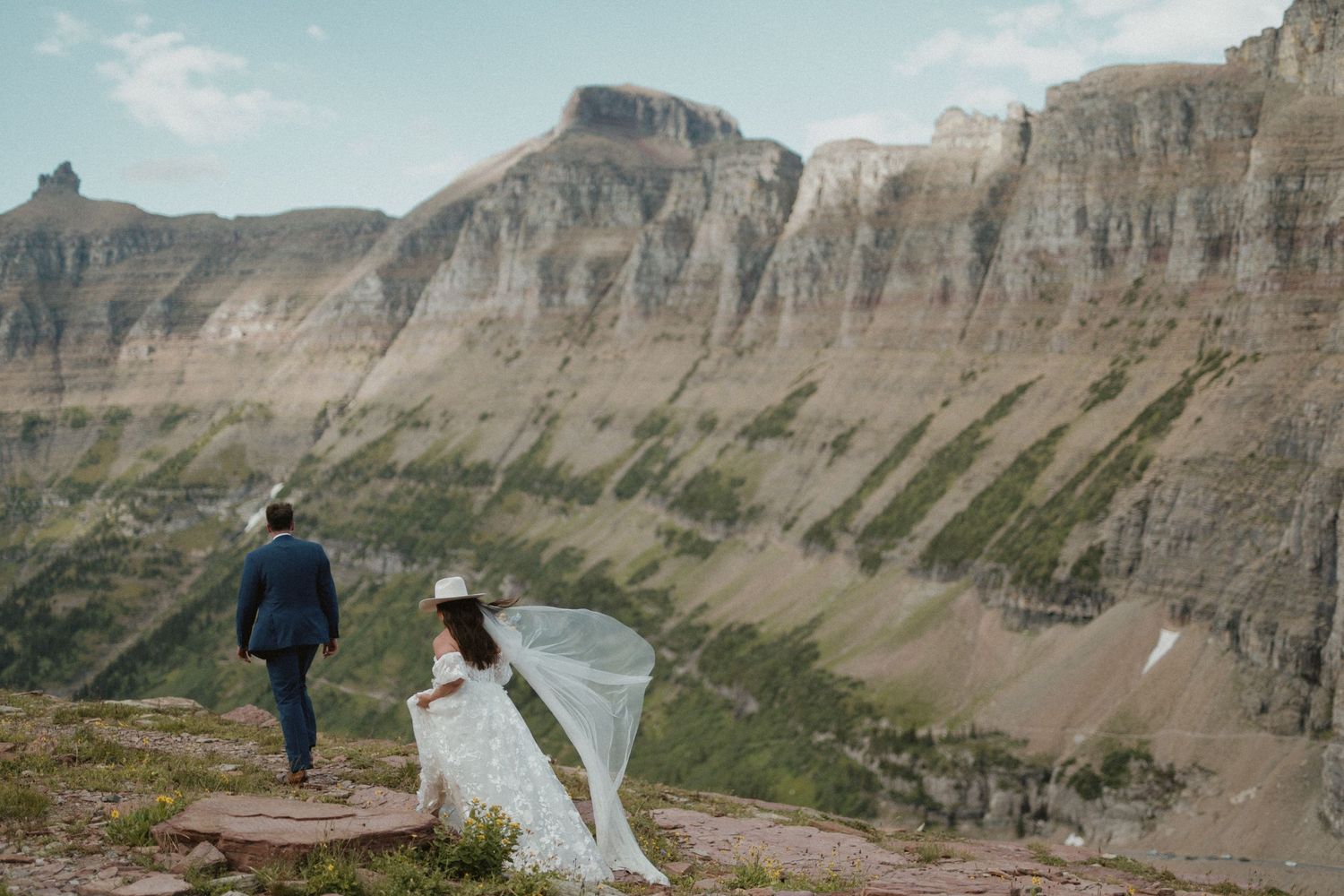 Bride and groom walk toward dramatic mountain cliffs during an outdoor elopement ceremony at Glacier National Park.