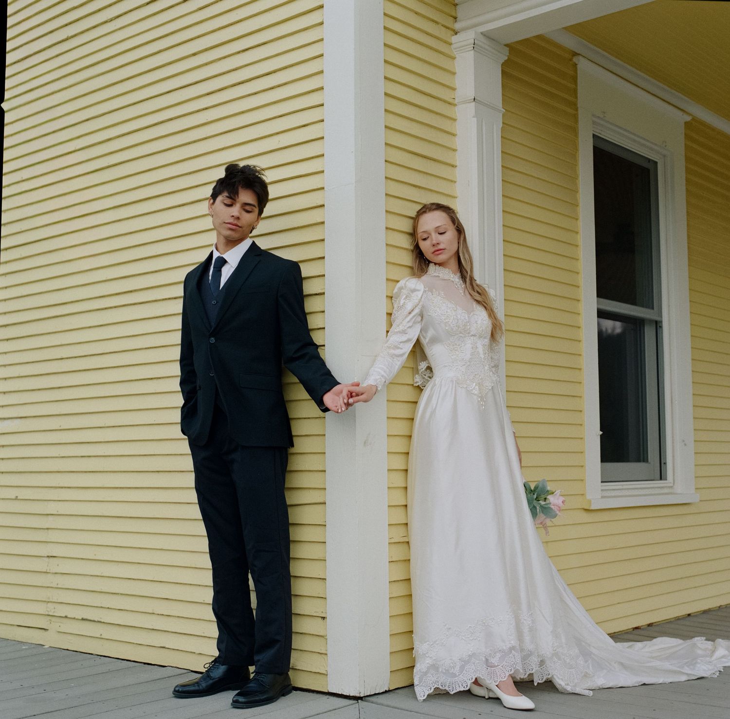 Wedding couple holding hands beside a yellow building, bride in lace gown and groom in black suit.