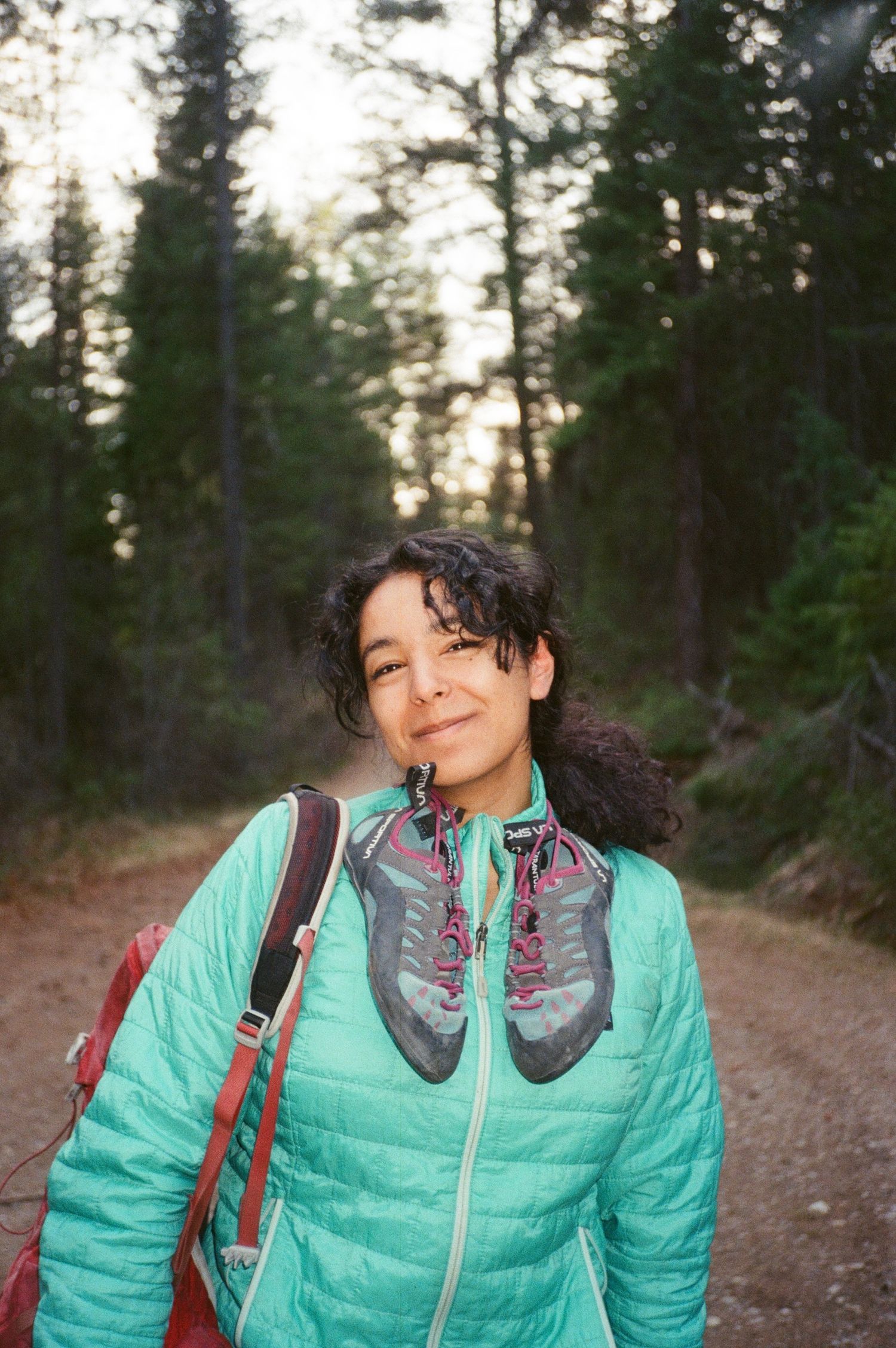 Woman in teal jacket with hiking shoes on chest, smiling on forest trail with red backpack.