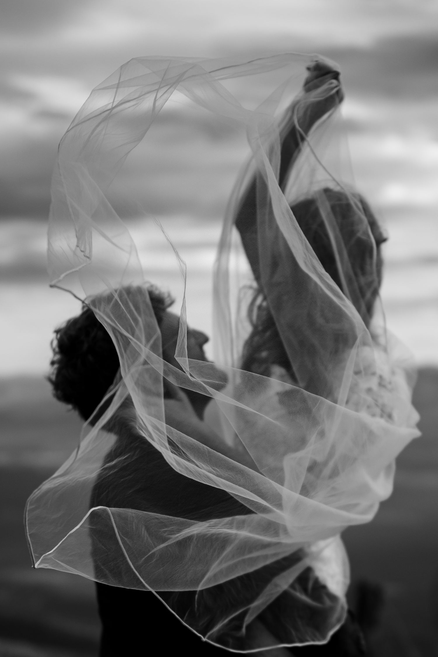 A dramatic black and white photo shows a flowing wedding veil billowing in the wind against a cloudy sky.