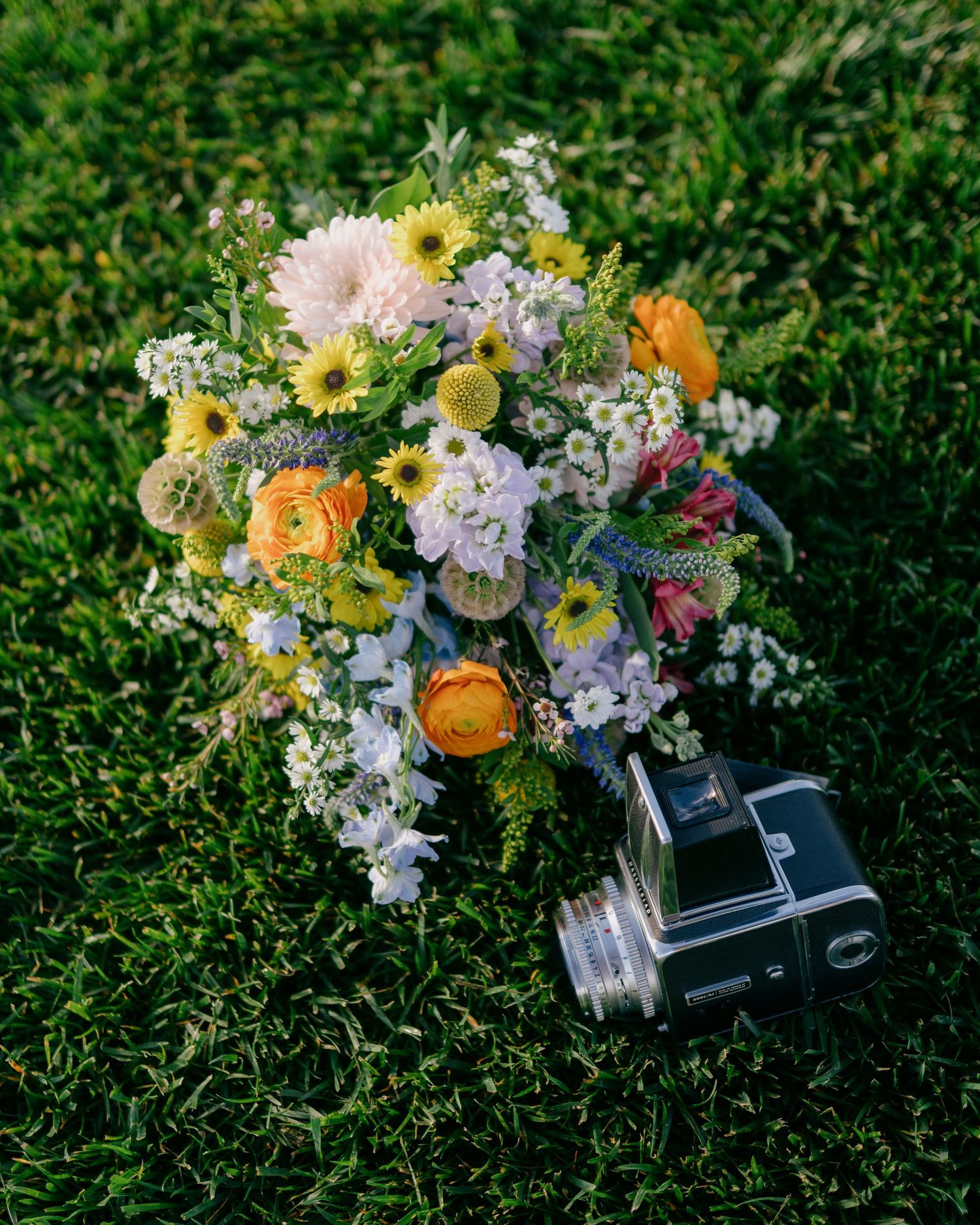 A bouquet of colorful flowers, including daisies and other blooms, rests on a grassy surface with a vintage camera nearby.