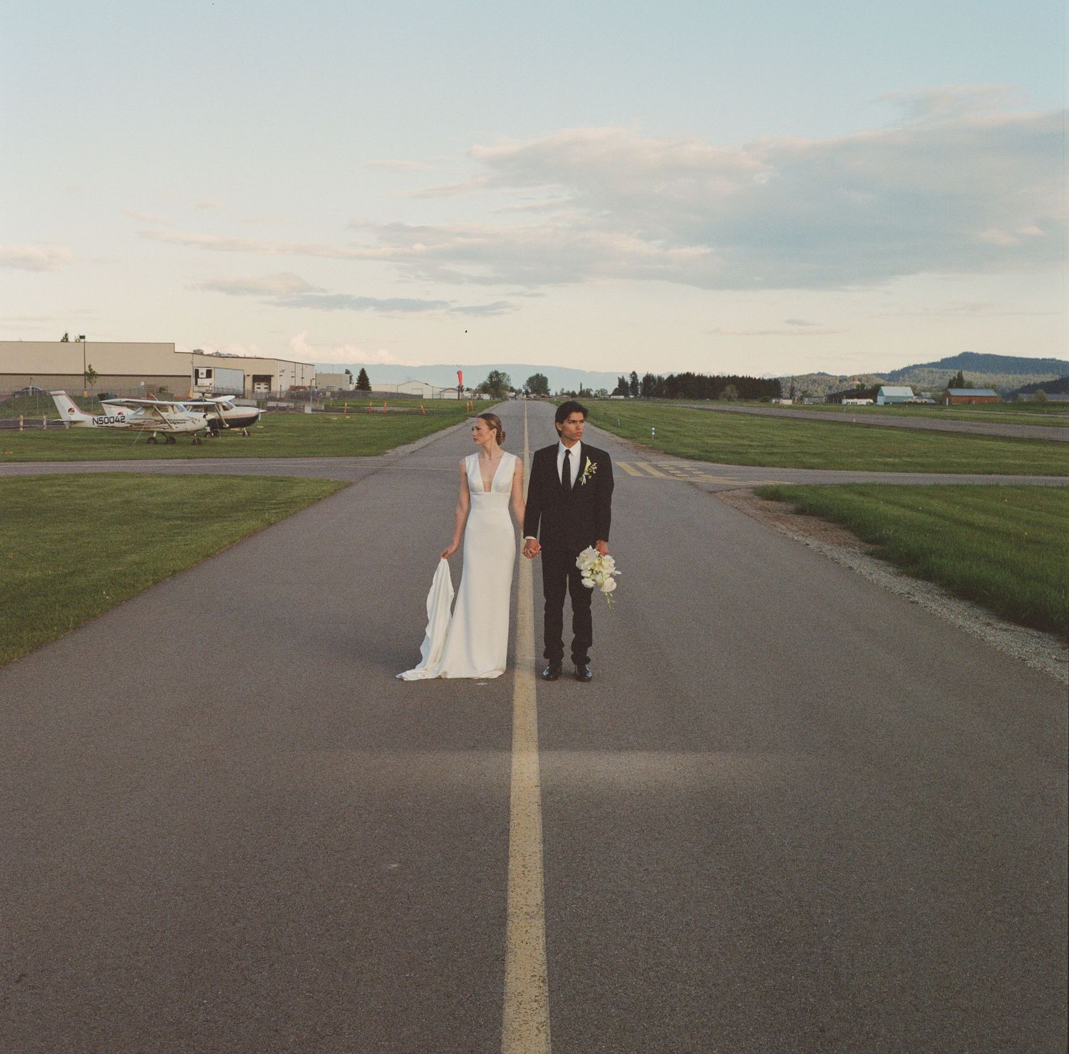 Bride and groom standing on airport runway in wedding attire, with planes and hangars visible in background.
