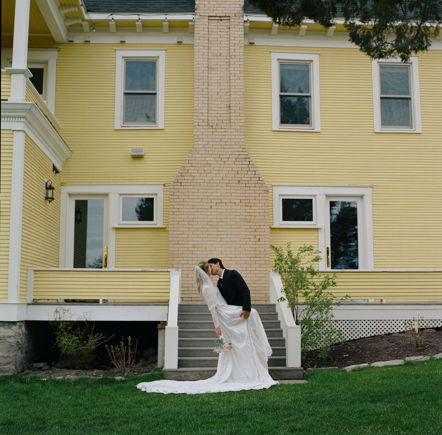 Bride and groom sharing a romantic dip kiss in front of a charming yellow Victorian house on their wedding day.