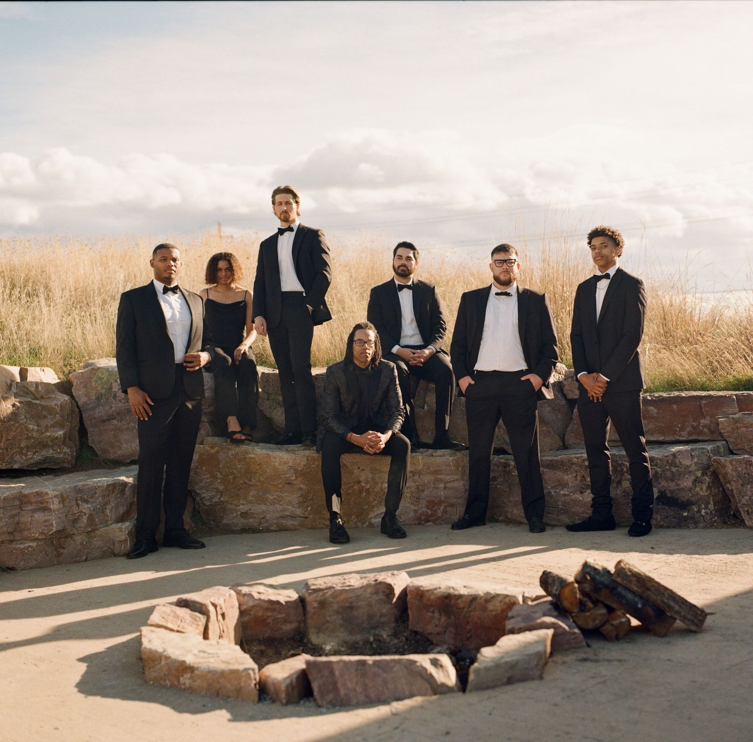 Formal wedding party relaxes on stone wall with scenic mountain backdrop.