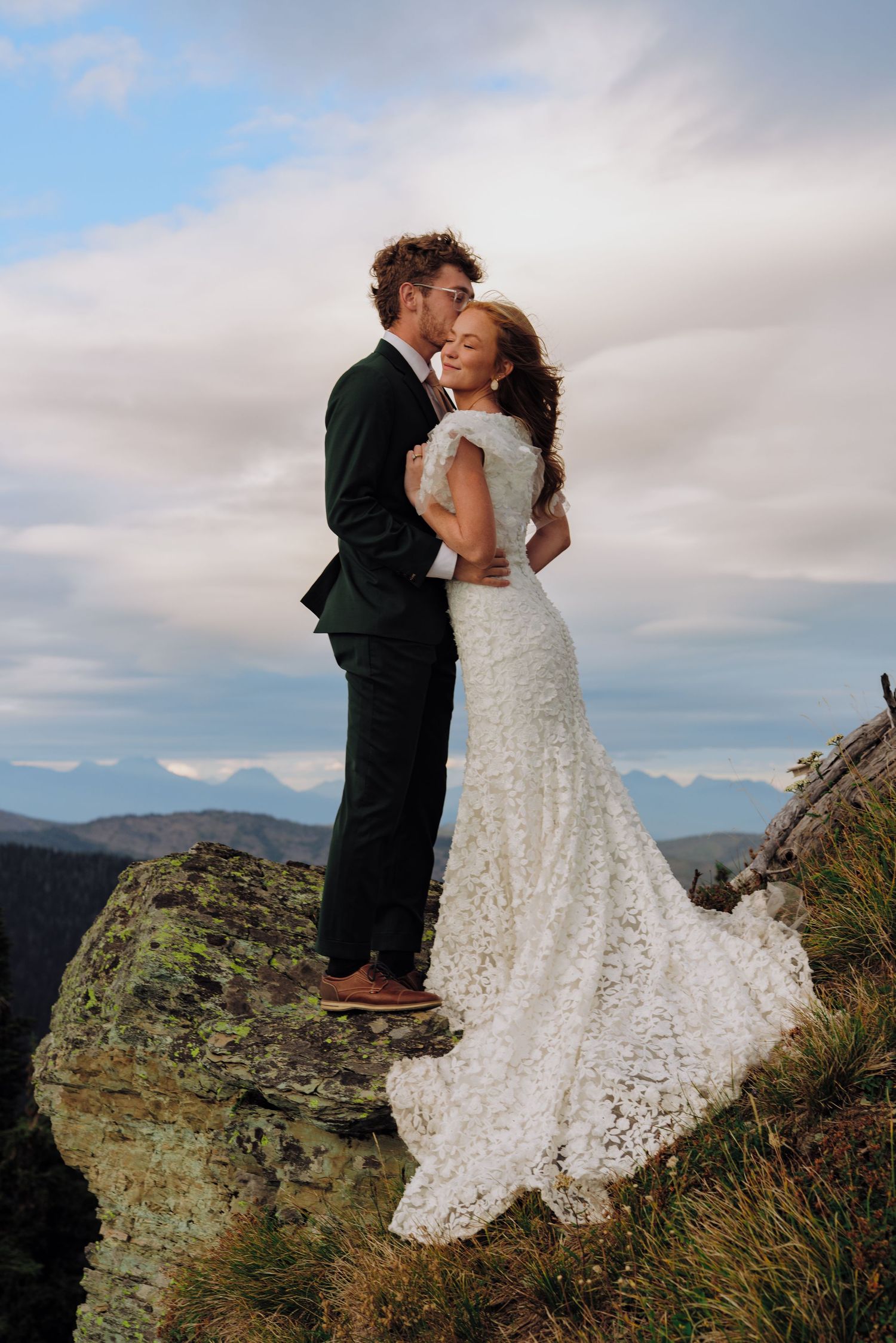 A romantic wedding couple embraces on a rocky mountain overlook with dramatic cloudy skies in the background.