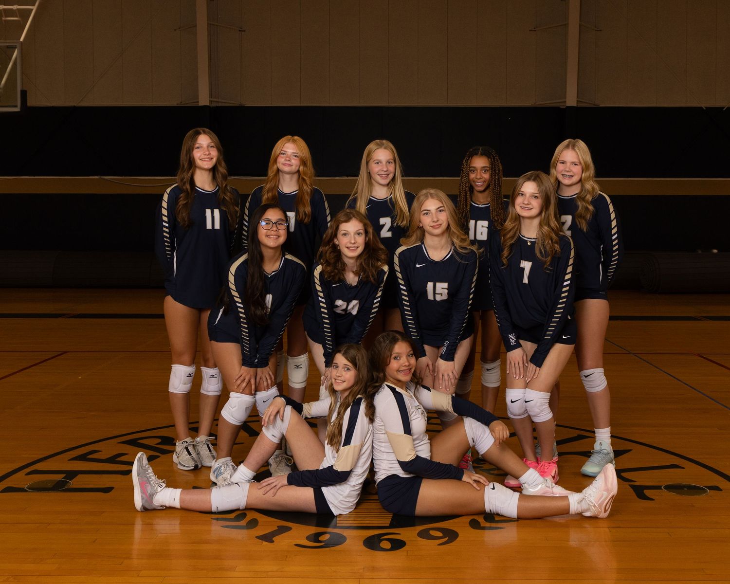 High school volleyball team members pose in navy uniforms on a wooden court floor for team photos.