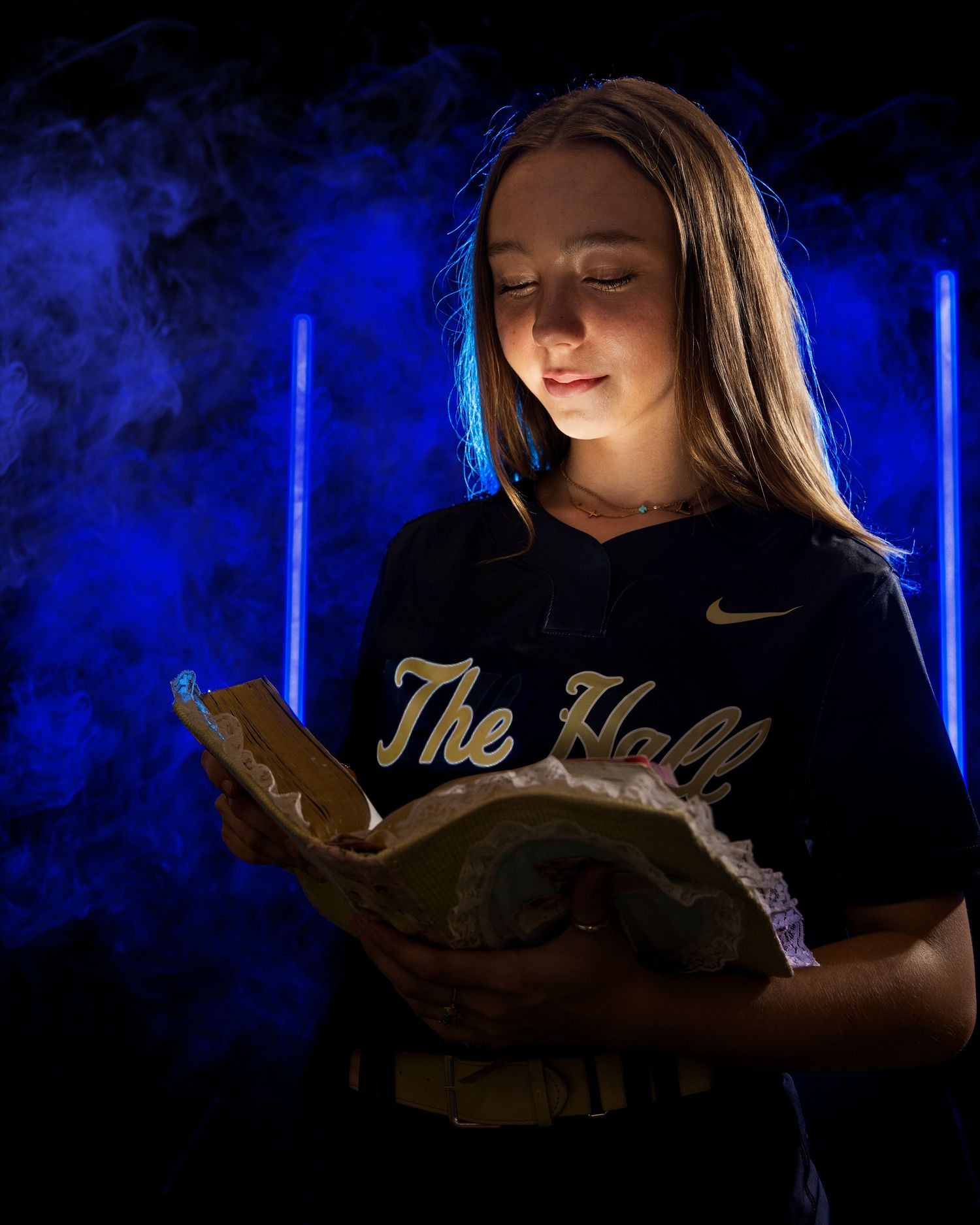 A person in a softball uniform reads from a bible illuminated by dramatic blue lighting and smoke effects.