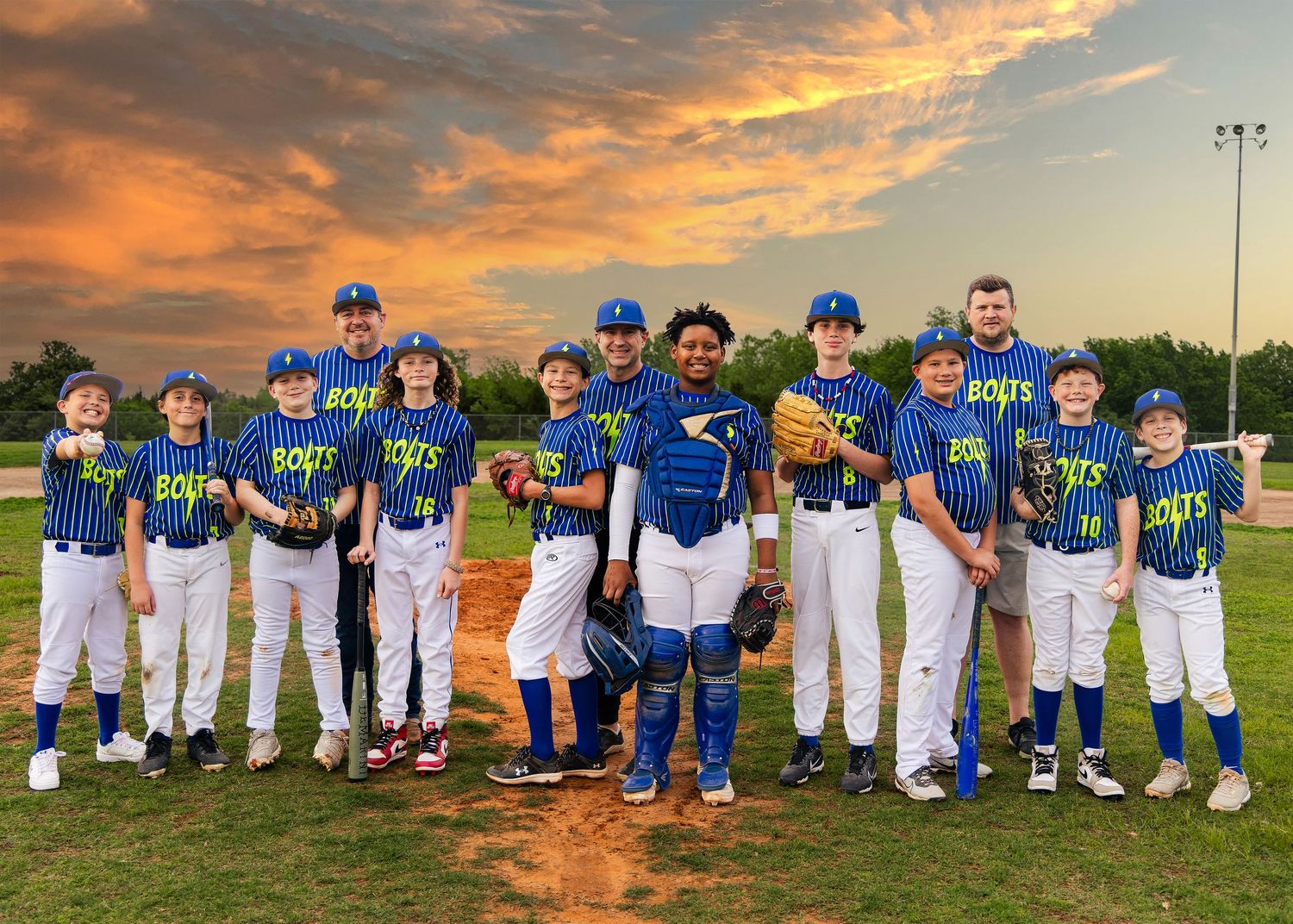 Youth baseball team players pose together on a field during sunset with vibrant orange sky in the background.