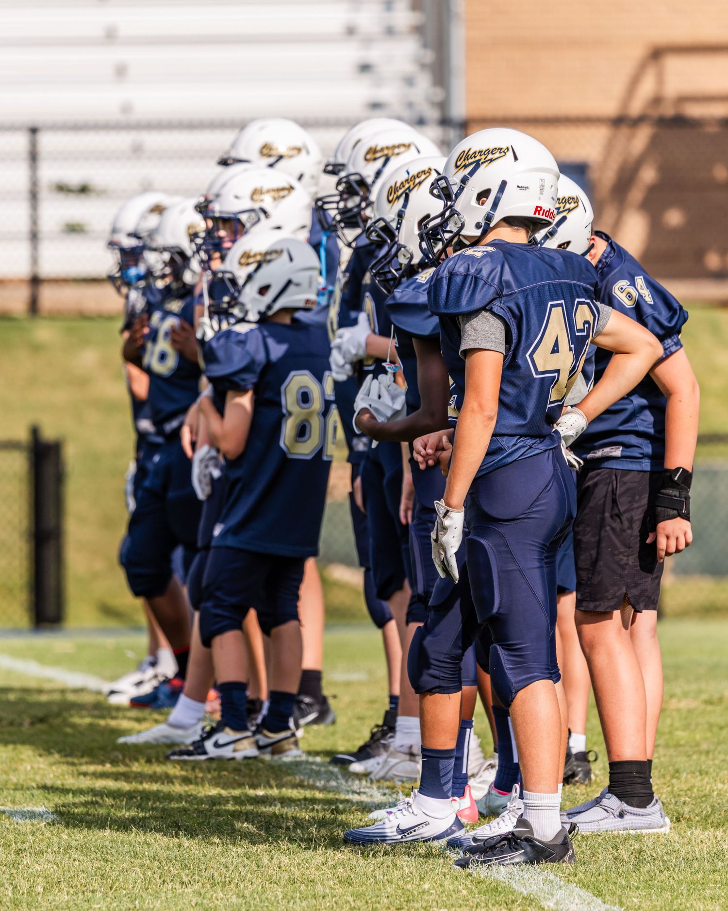 Football players in navy blue uniforms kneel in a line on a grass field during practice or pre-game preparation.