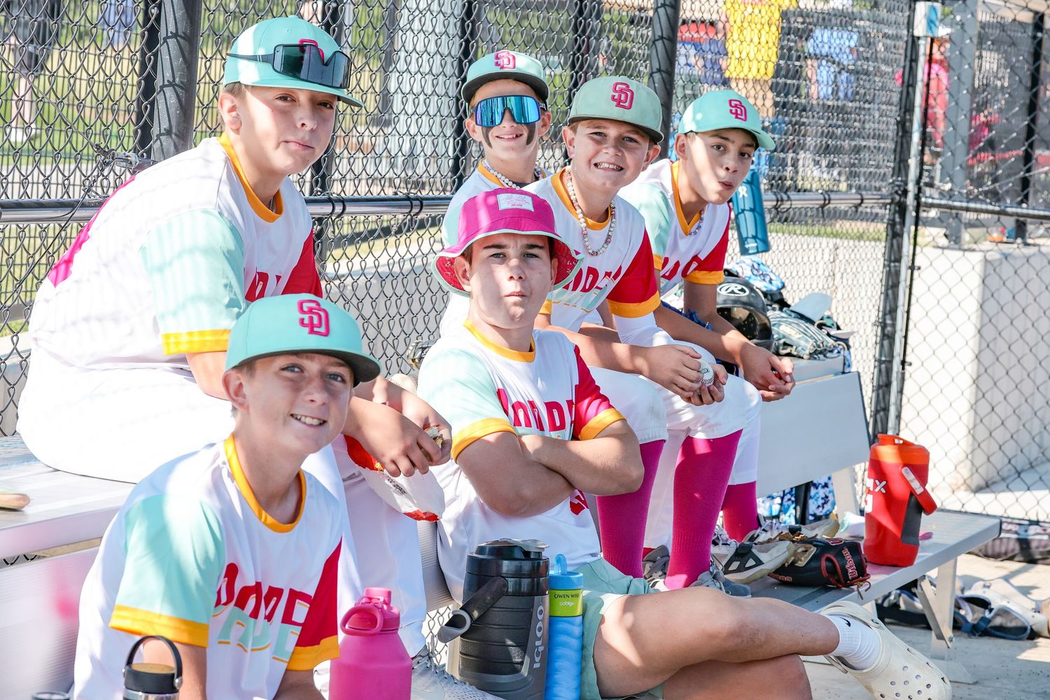 Youth baseball players in colorful uniforms sit together in a dugout during a game.