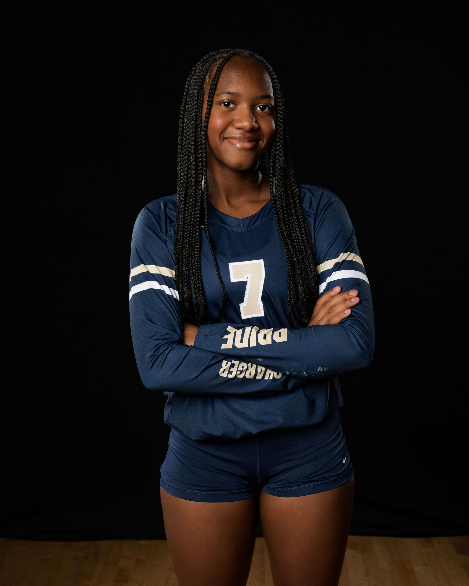 Athletic figure in navy blue uniform with long braids poses with crossed arms against dark background.