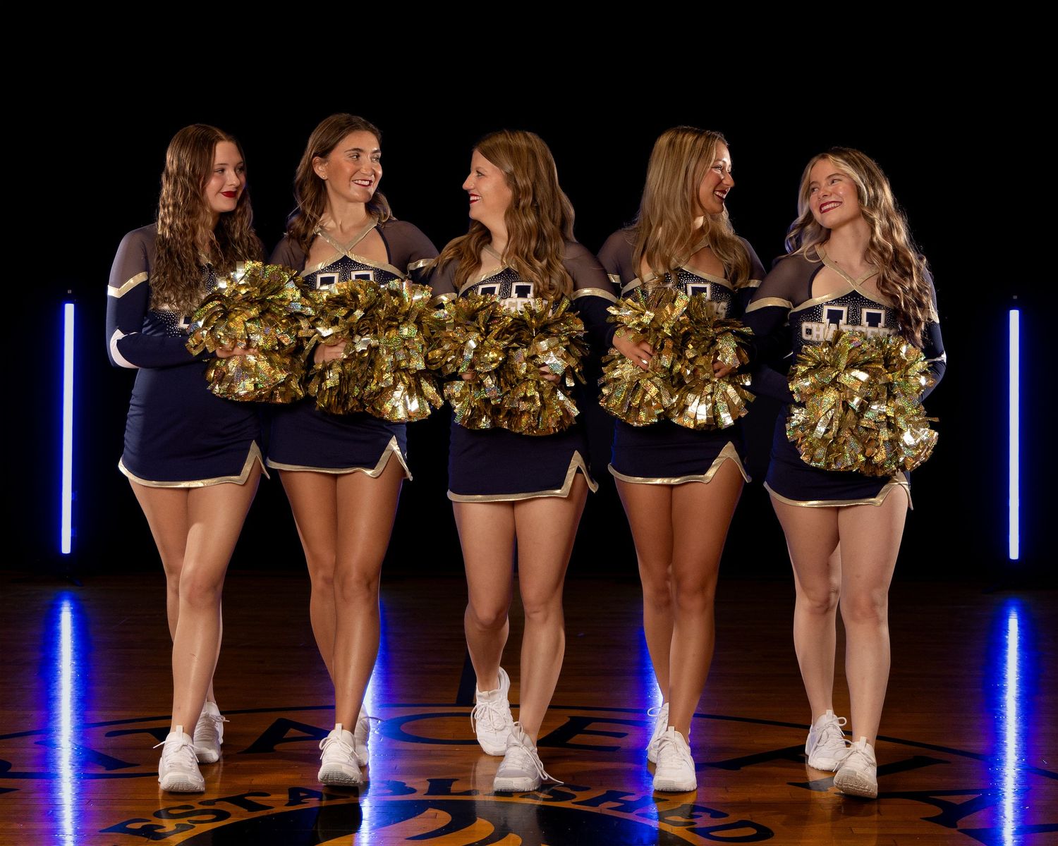 Cheerleaders in navy uniforms with gold pom-poms pose together on a dark stage with blue lighting effects.