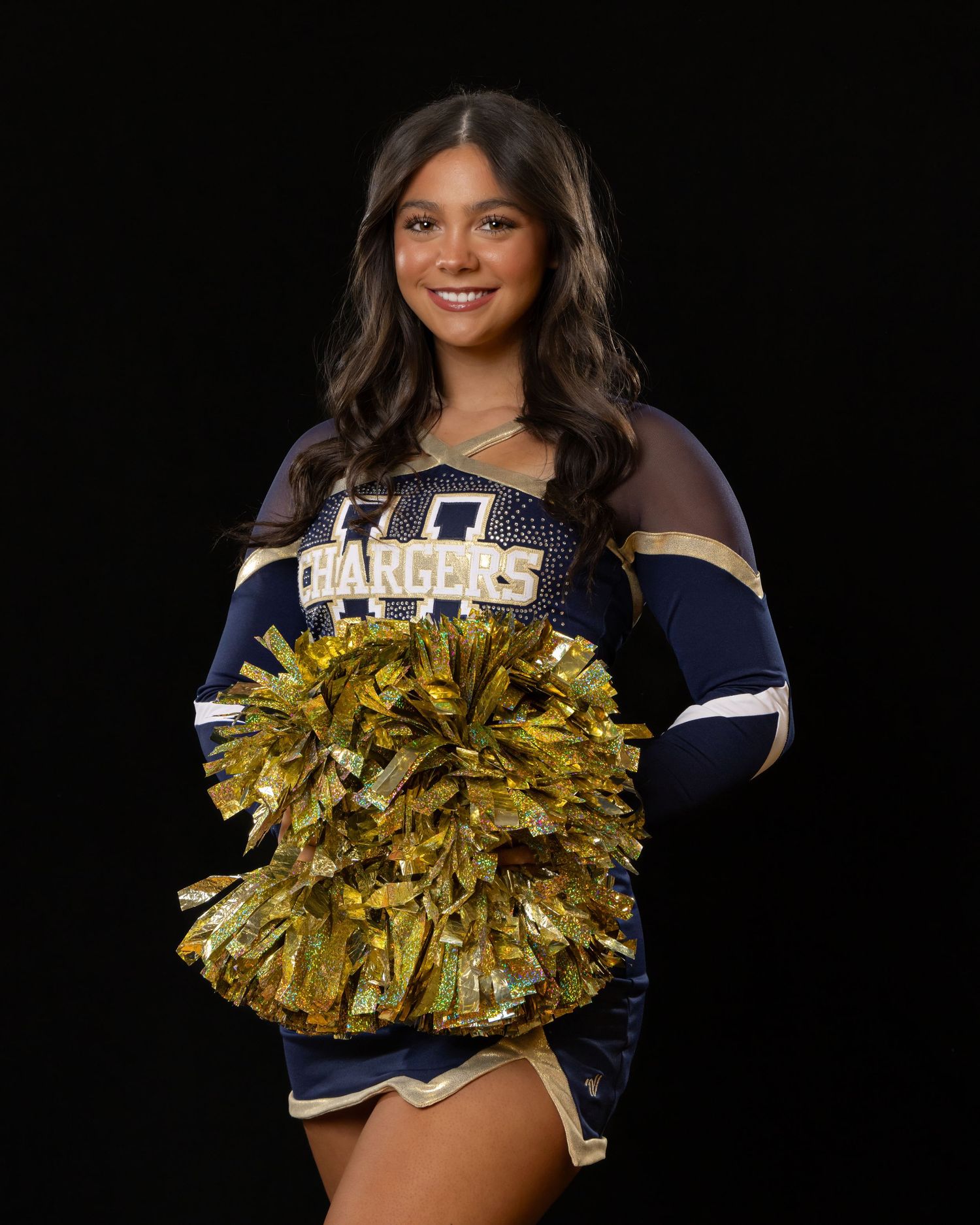 cheerleader in navy and white uniform with gold pom poms performs against black background.