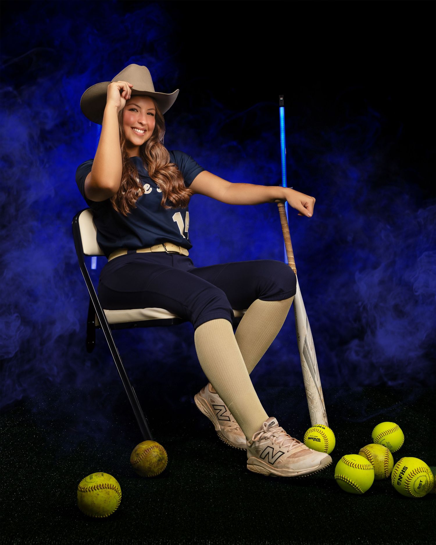 Softball player in navy uniform and cowboy hat poses with equipment against dark background with blue smoke effects.