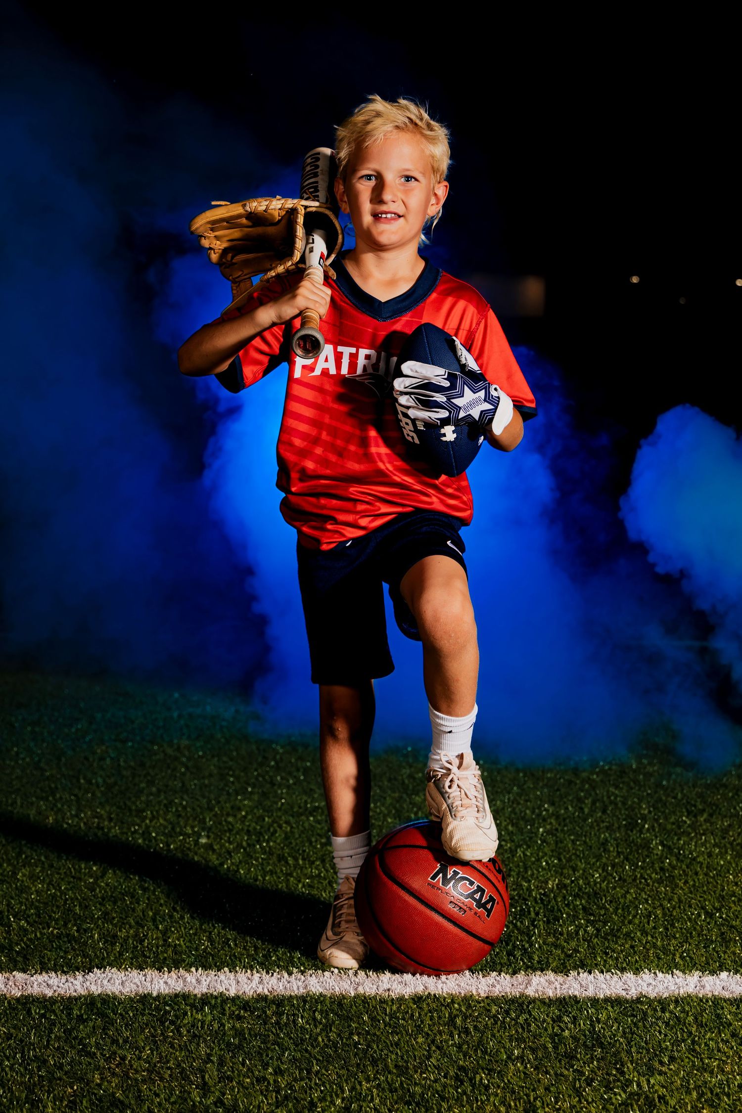 Young flag football player in red uniform poses with ball on field at night with dramatic blue lighting effect. Multi-sport player