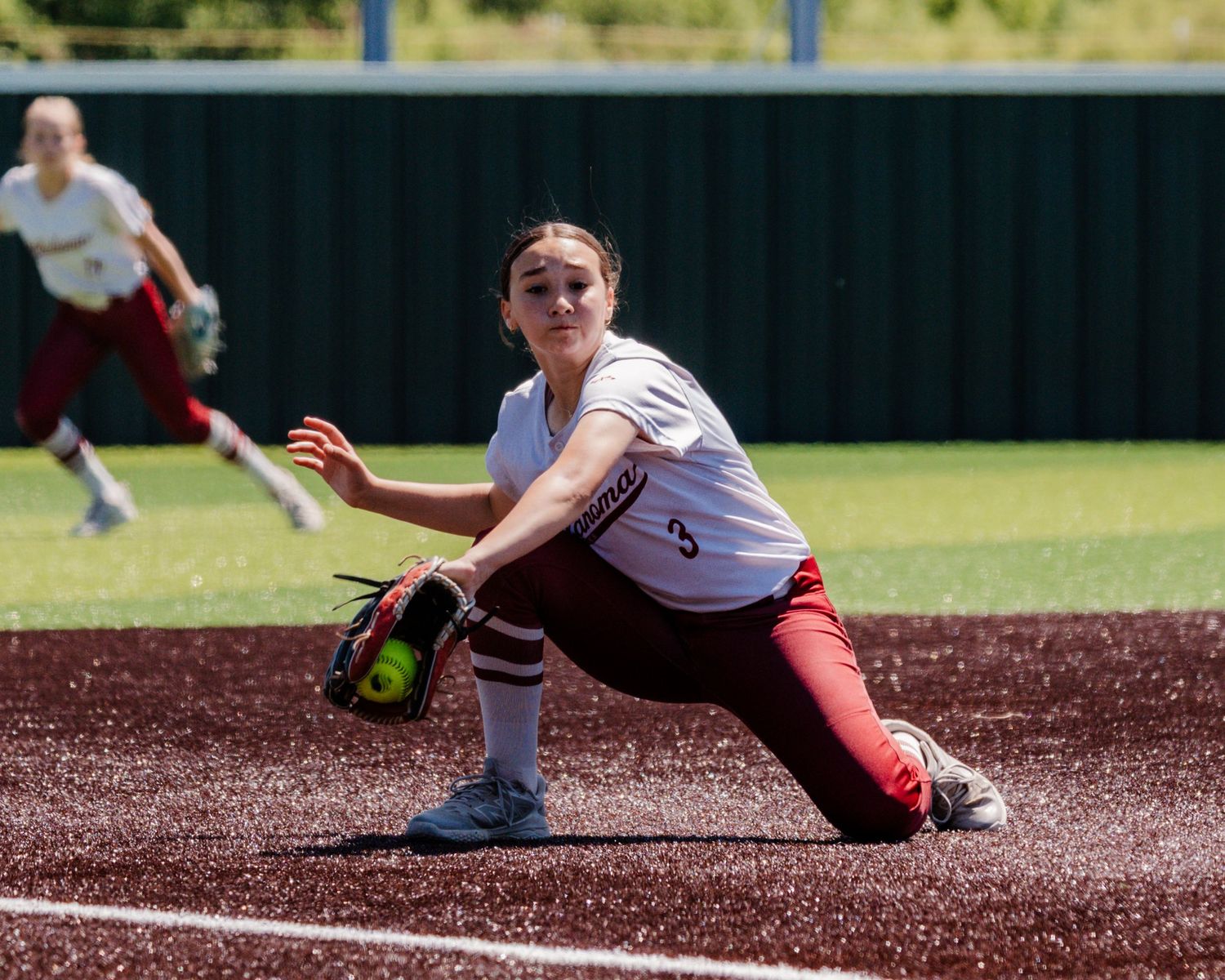 Softball player in white jersey and red pants stretches low to field a ground ball during a game.