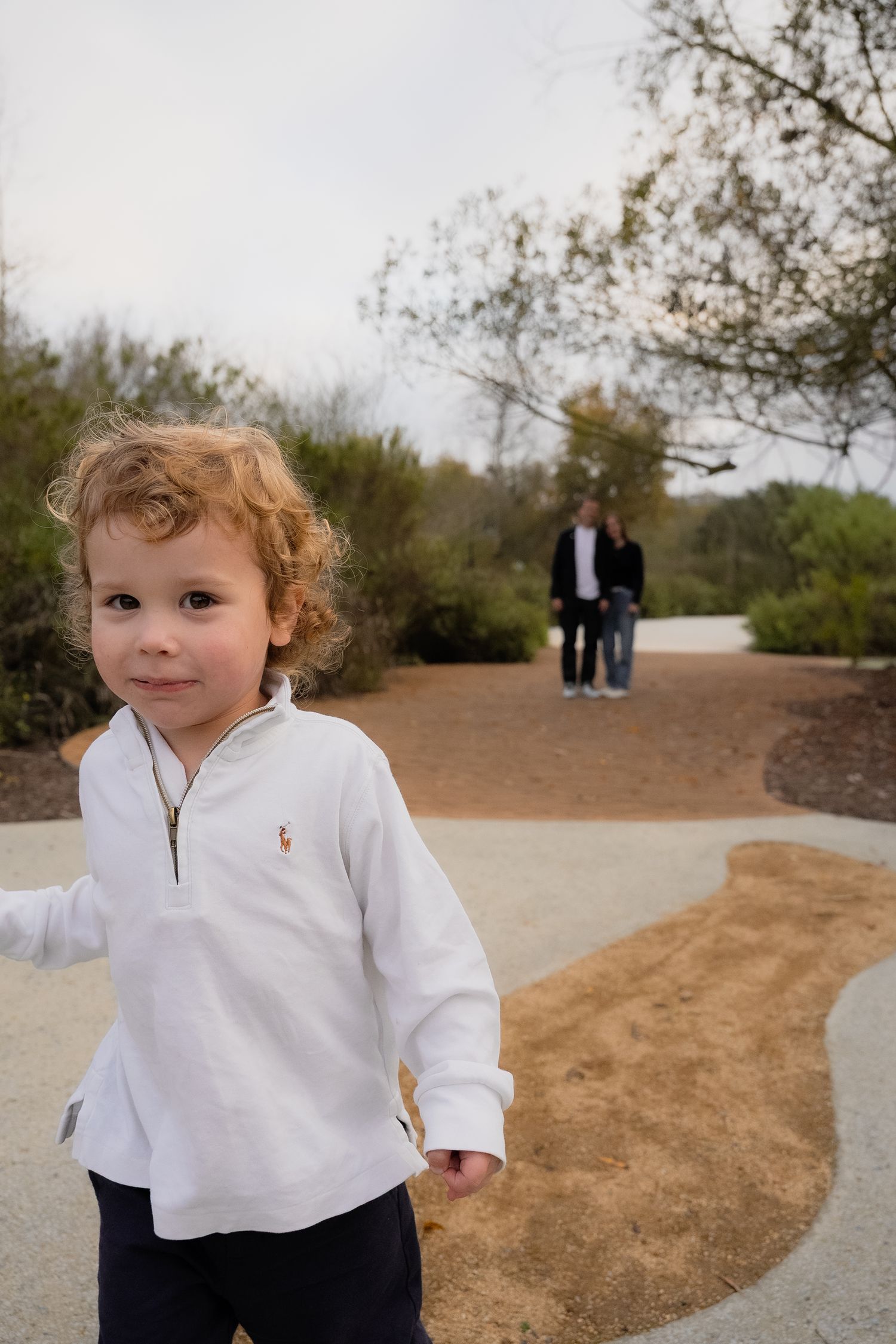 a natural portrait of a toddler boy smiling and walking towards camera at the park while his parents look on in the background
