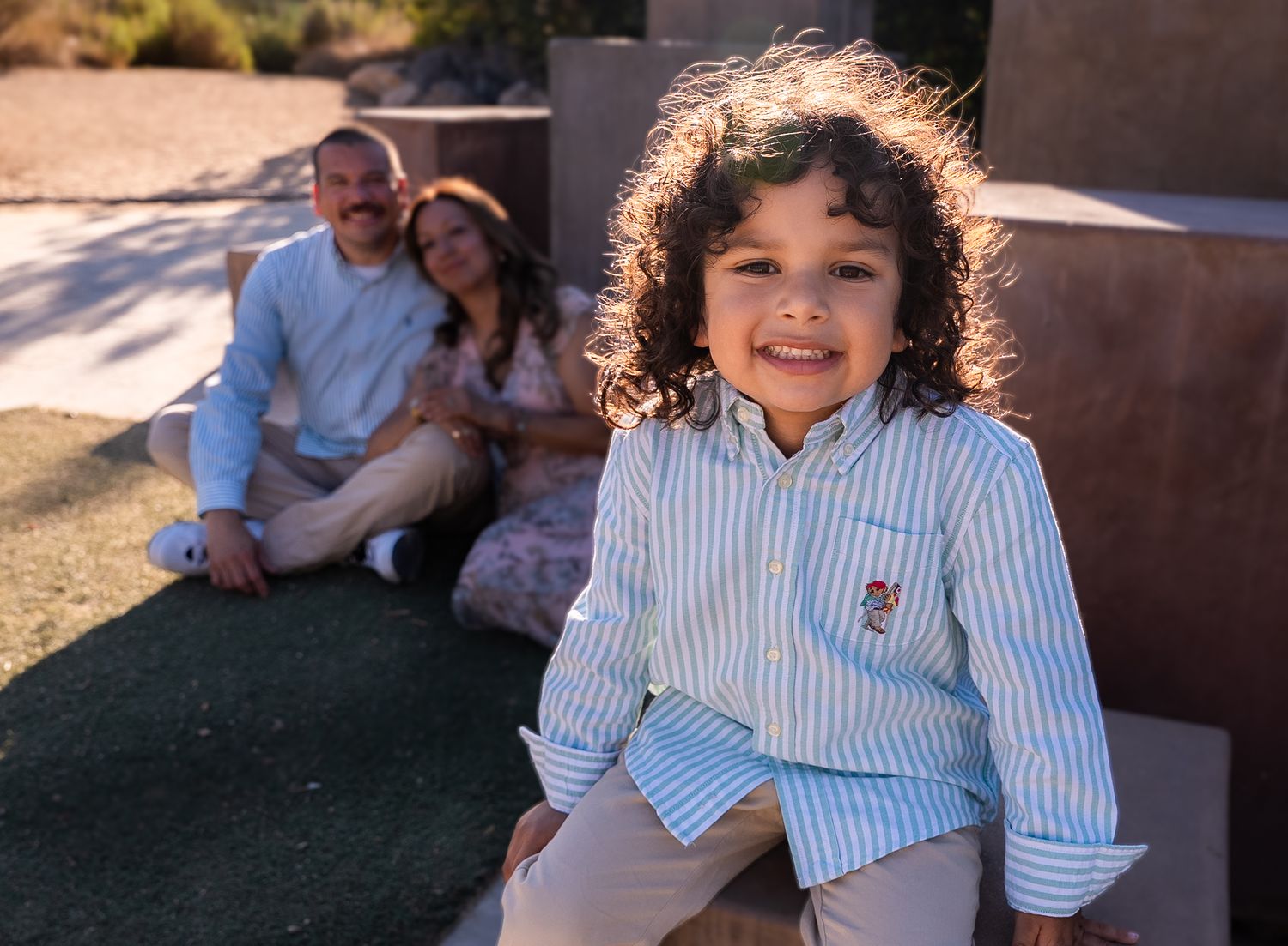 a relaxed portrait of a four year old boy with shoulder-length curly hair smiling at the camera with his parents looking proudly on