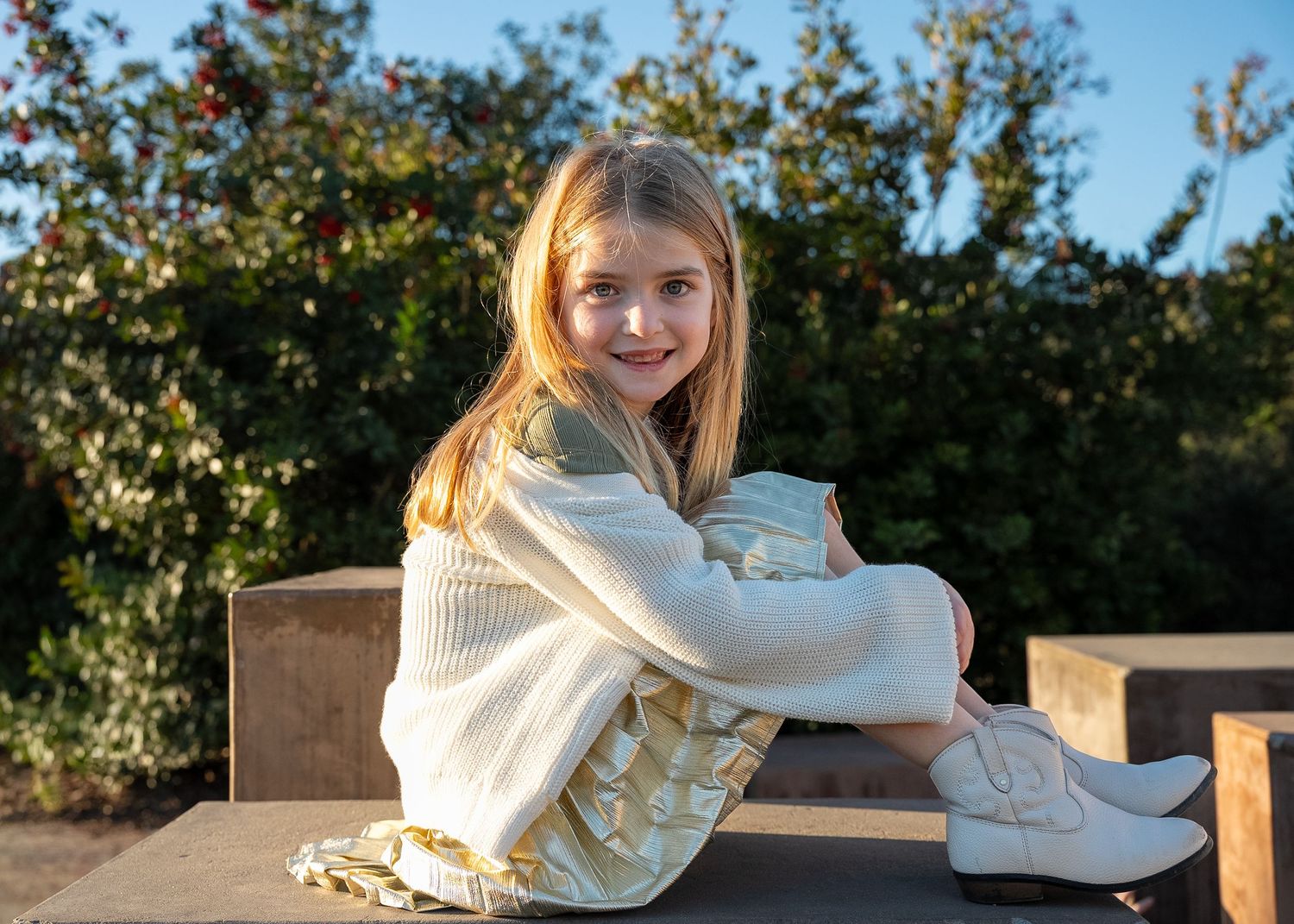 a portrait of a seven year old girl holding her knees while she smiles at the camera during golden hour