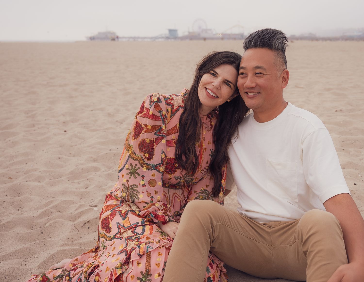 a natural relaxed portrait of a couple sitting in the sand on the beach with the pier in the background