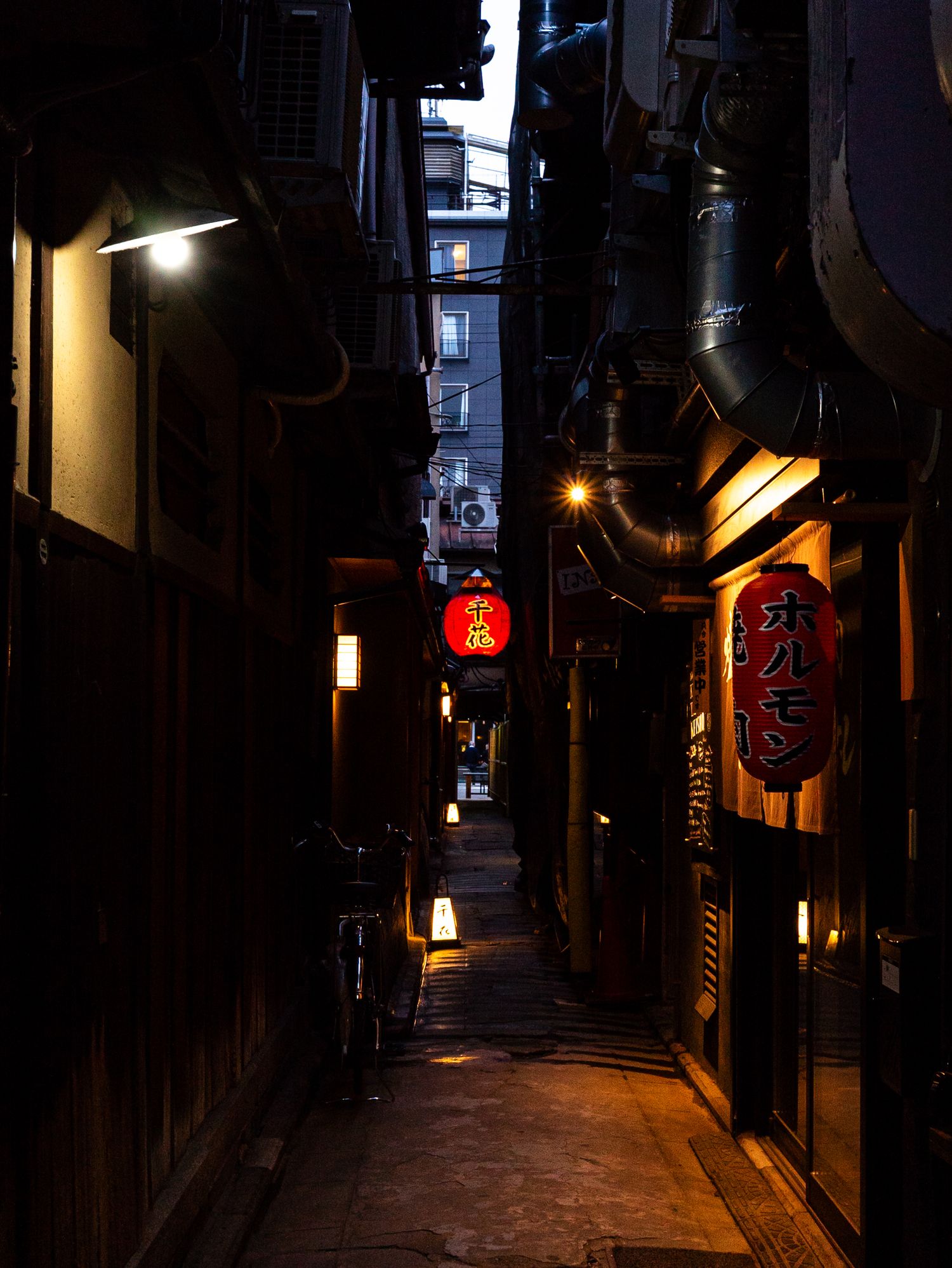 A dark and narrow historic Kyoto alley that ends in a sliver of a well-lit modern building in the center of the photo
