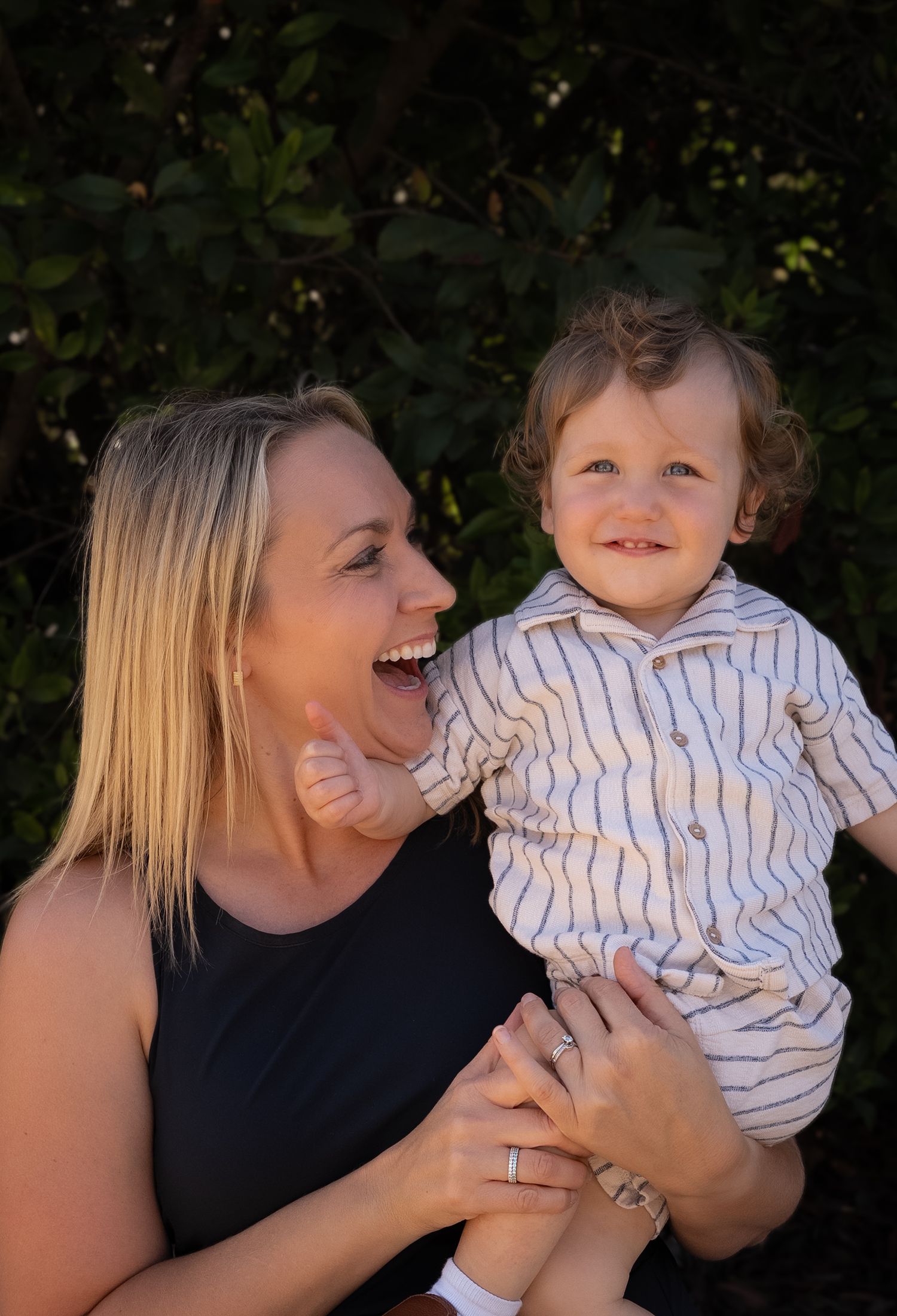 A mother holding, laughing and looking at her toddler boy while he smiles at the camera