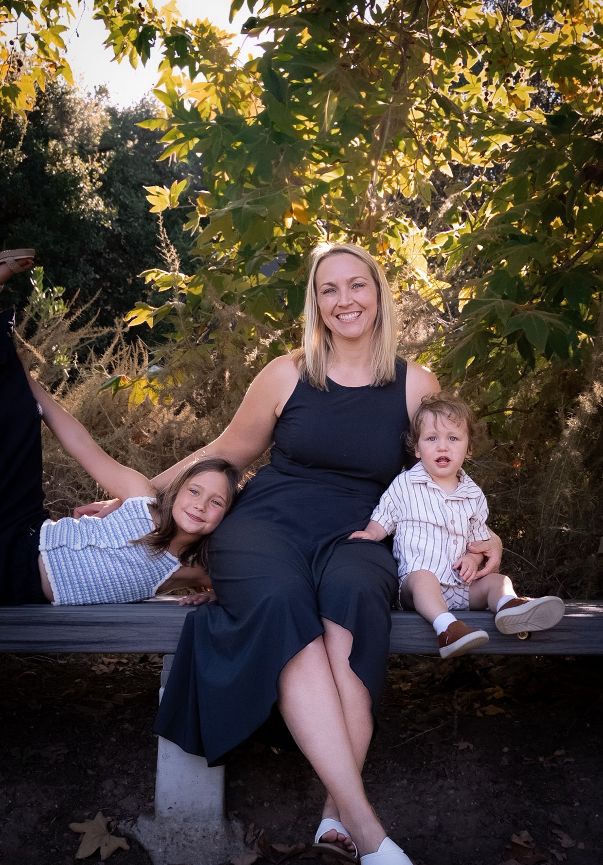 a photo of a mother and her toddler and seven year old daughter on a bench at the park