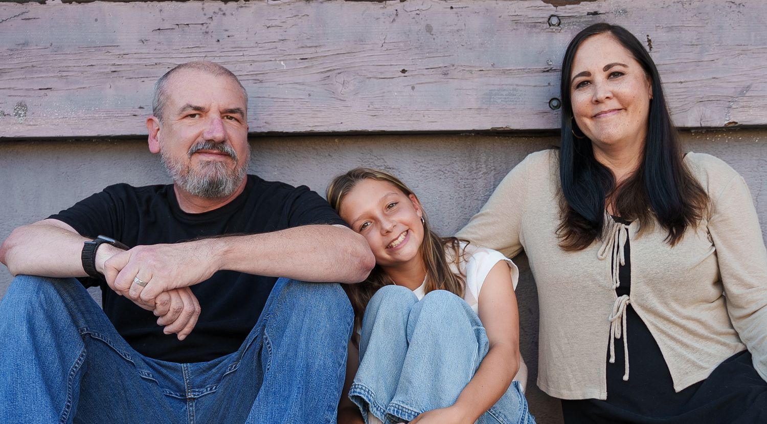 a portrait of a family of three sitting against a faded pink wall smiling at the camera