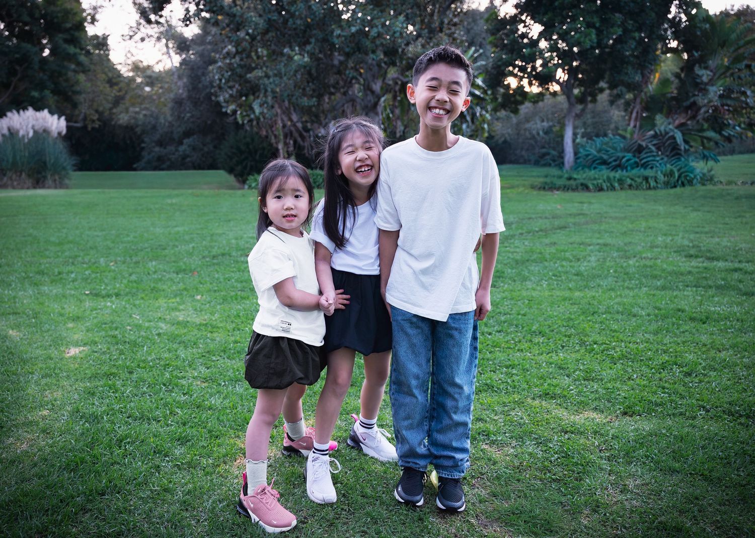a loving photo of three siblings holding hands and hugging at the park while smiling at the camera