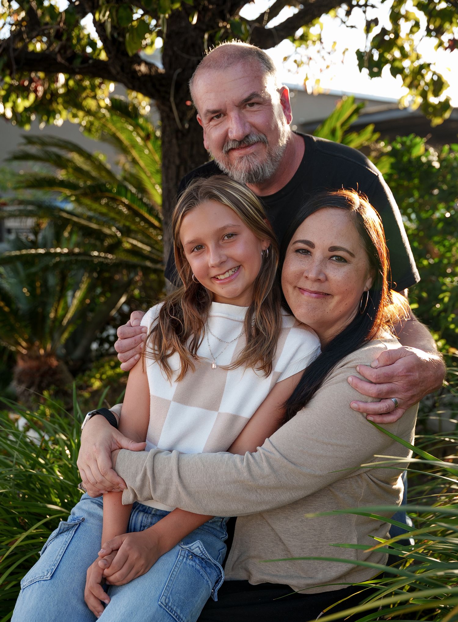 a portrait of a family of three smiing at the camera in front of greenery