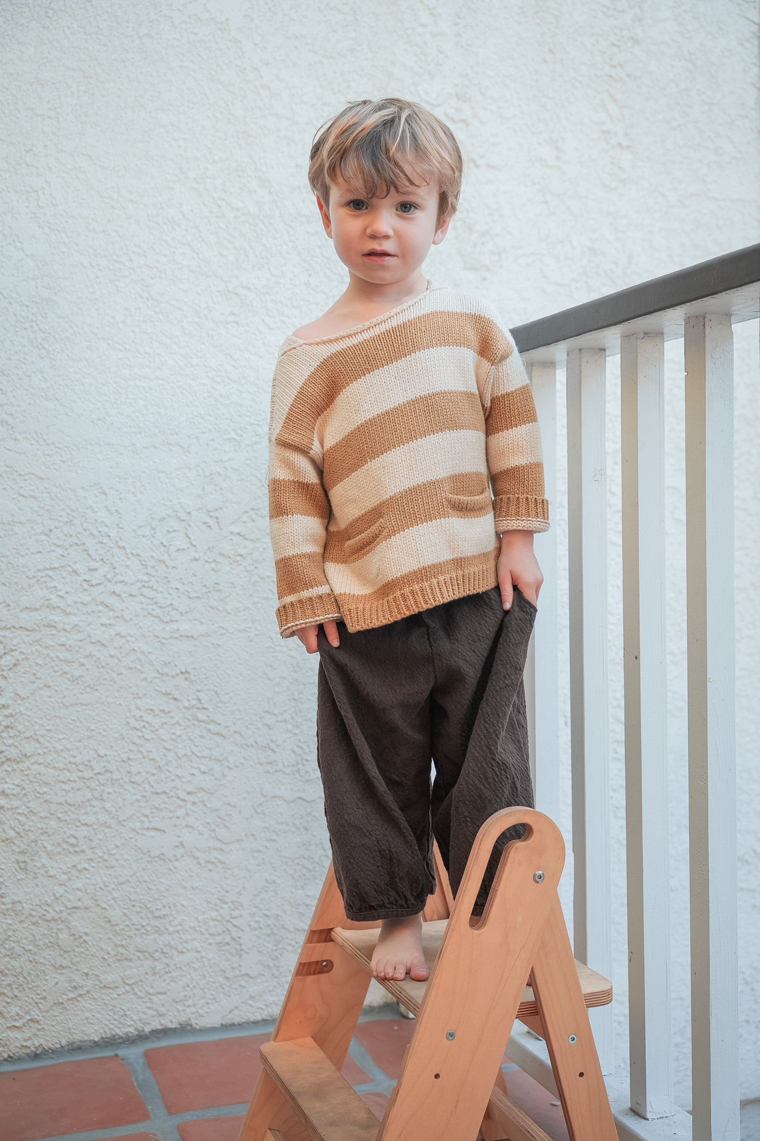 a toddler in a striped sweater standing on a stepstool looking at the camera