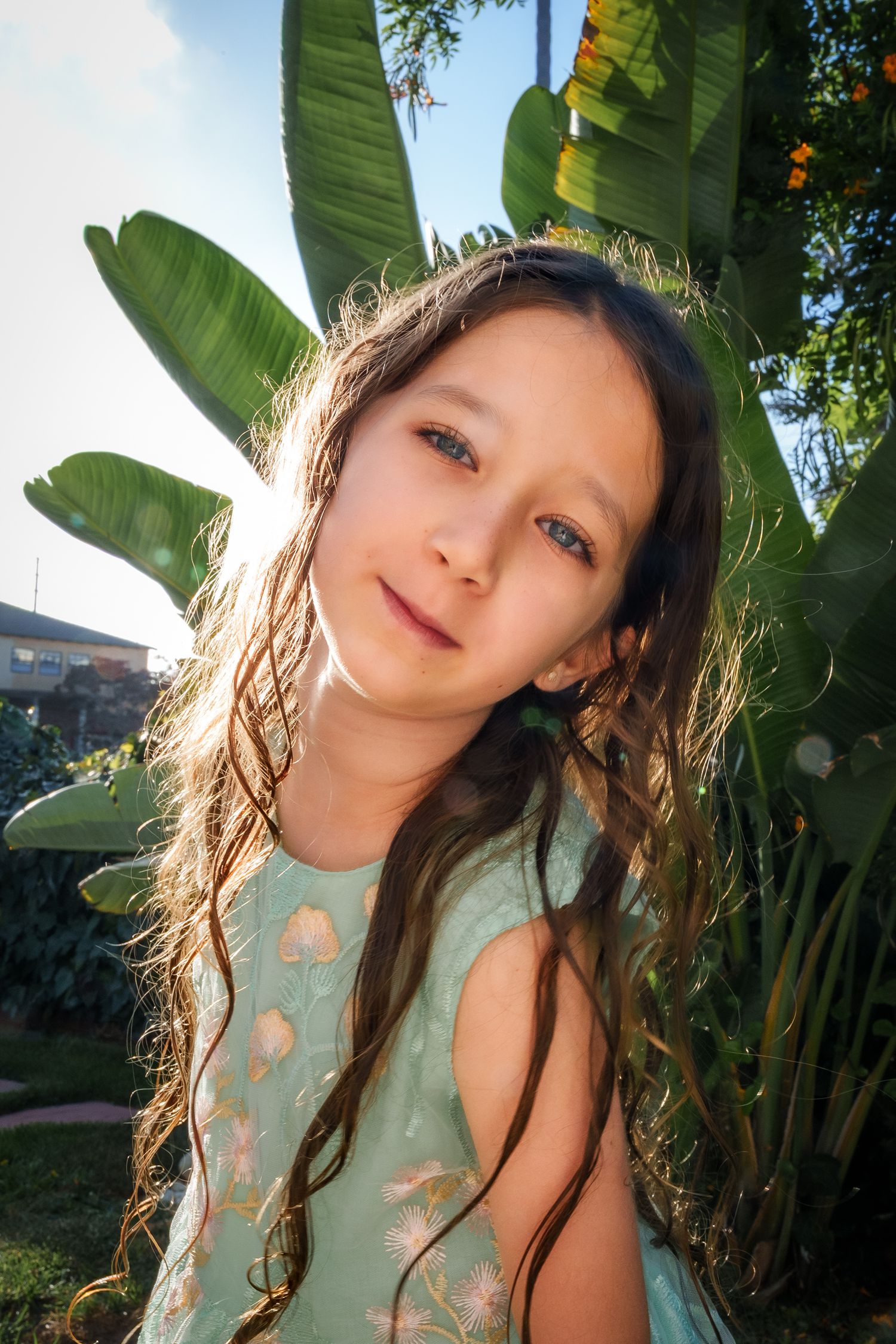 a seven year old girl with long hair looking at the camera with a large banana leaf plant in the background