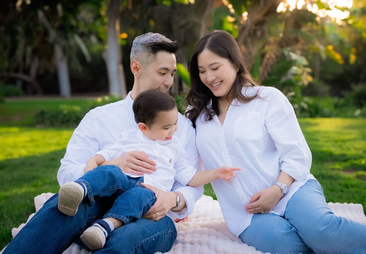 a toddler pointing at his mother's pregnant belly as they all relax on a blanket together at the park