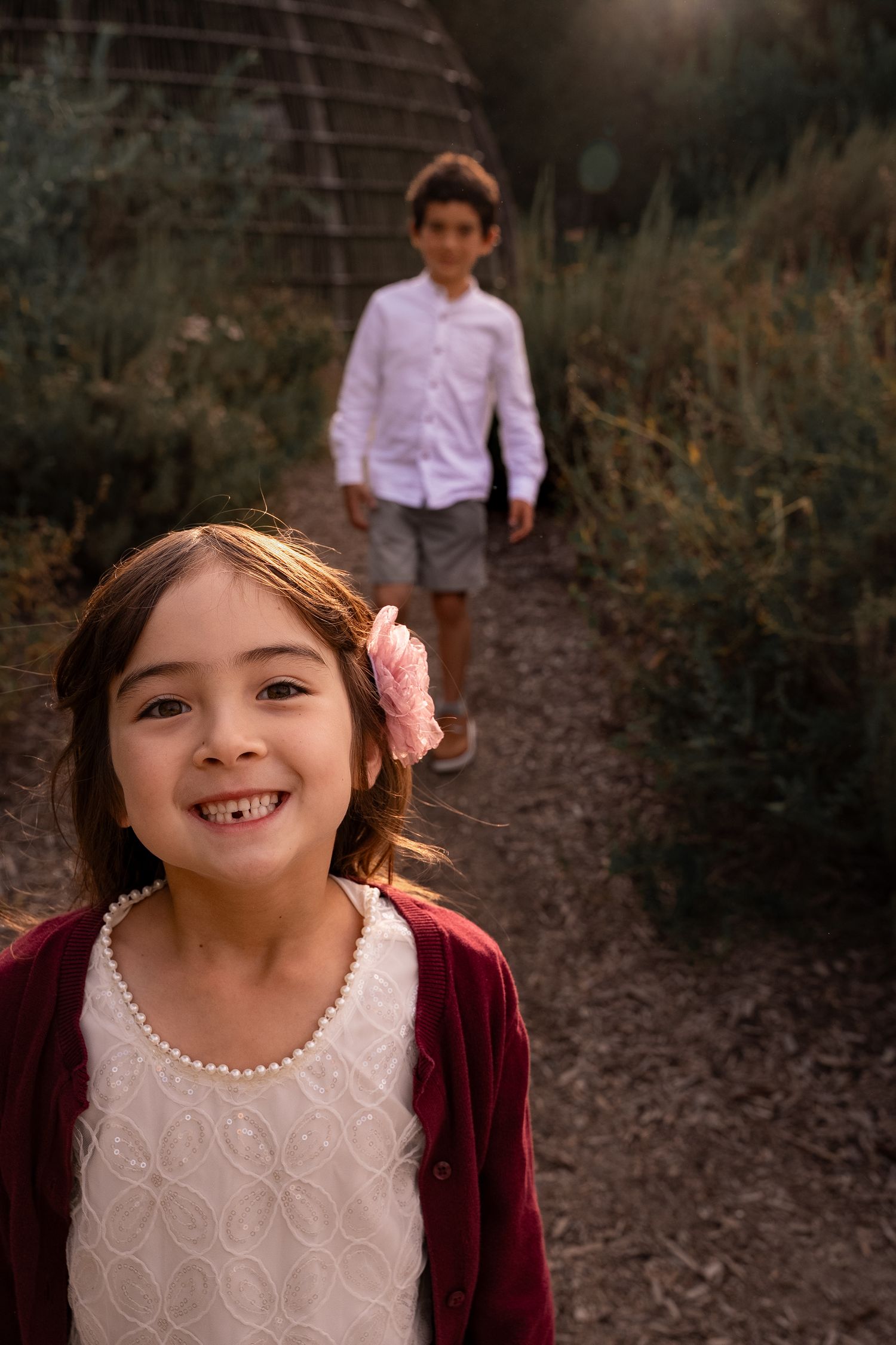 a smiling portrait of a girl in a pretty dress at the park smiling while her brother walks towards her in the background